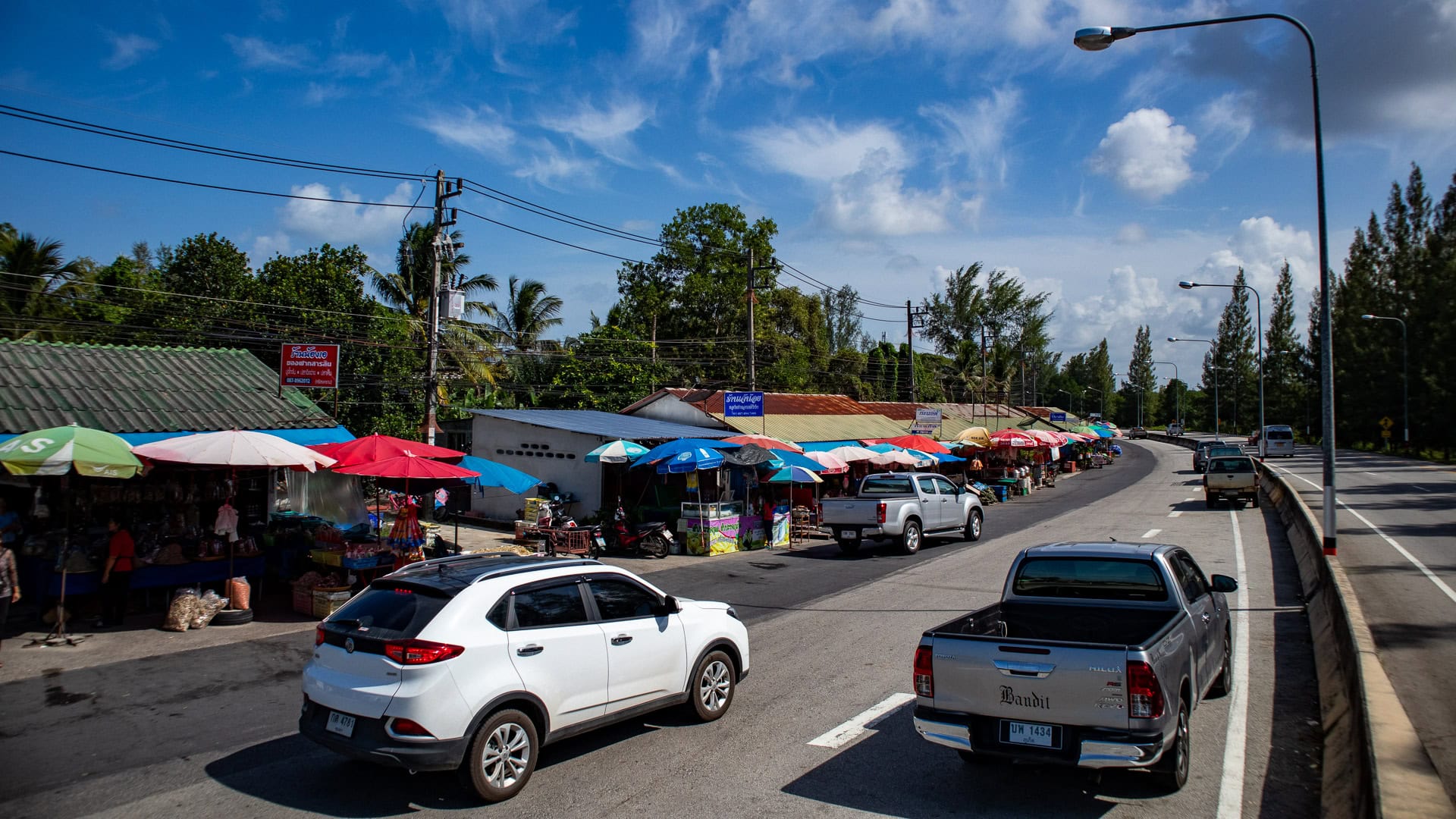 Roadside dried fish market along the Sarasin Bridge in Phuket with colorful umbrellas, parked cars, and stalls set up beside a busy road.