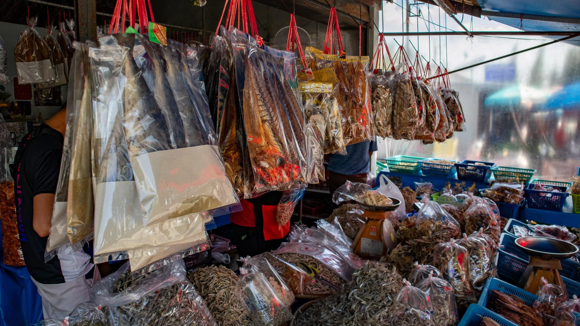 Hanging bags of dried fish and seafood displayed inside a roadside market stall, with baskets and scales visible behind.