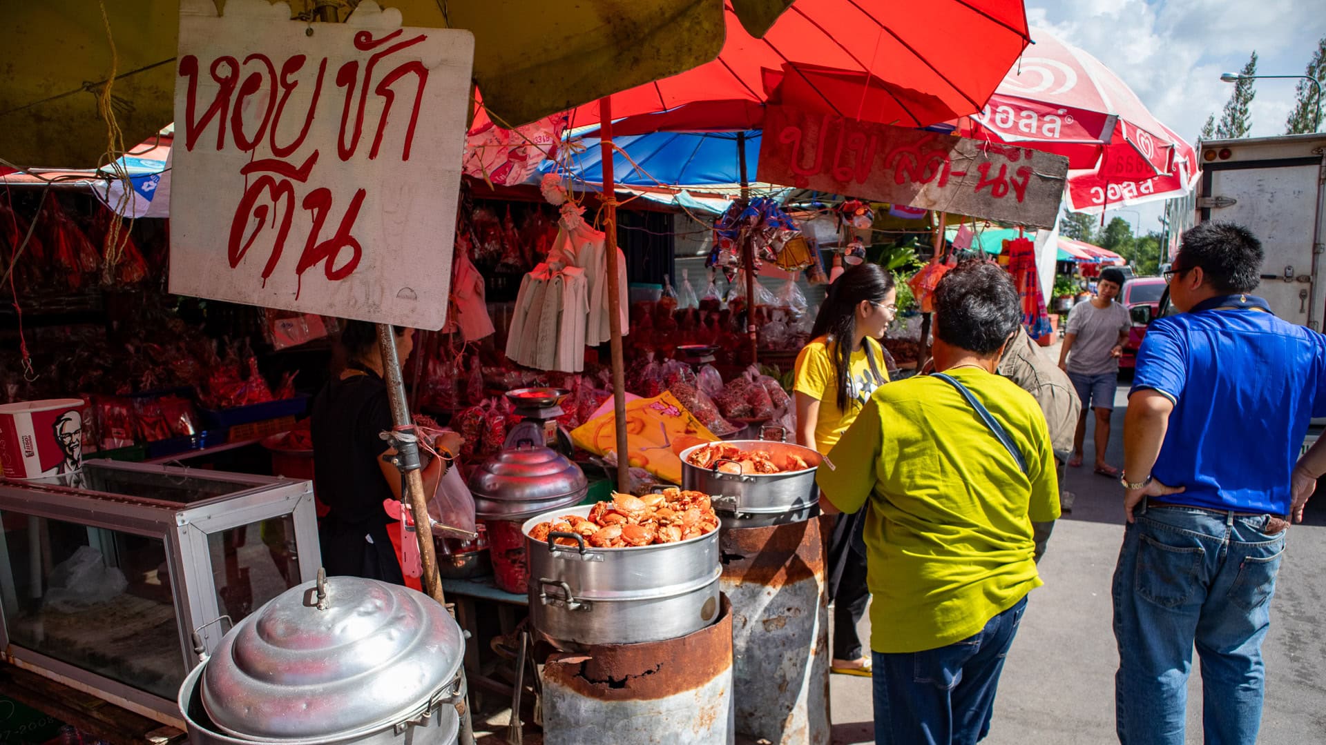 Customers buying food at a roadside dried fish stall, with steaming pots, umbrellas, and traffic passing close by.