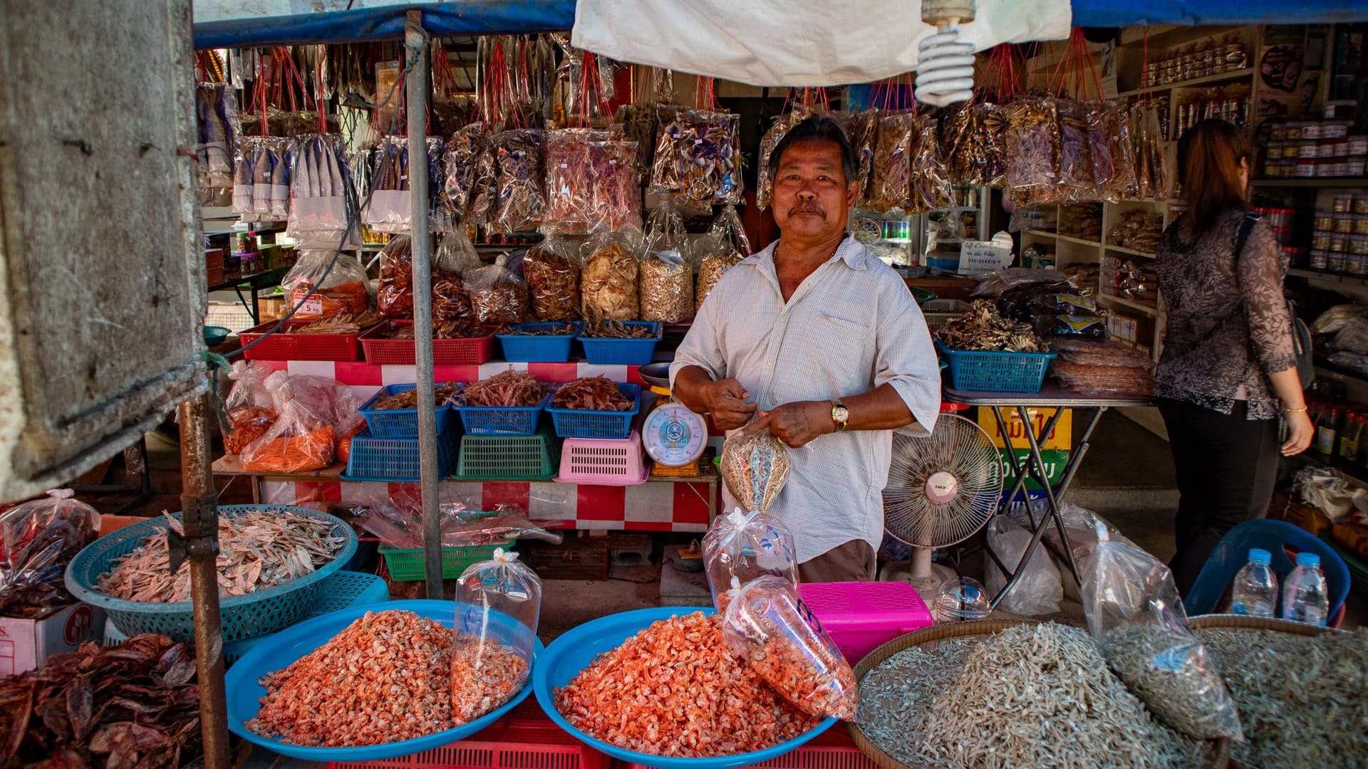 Vendor standing behind a dried seafood stall with baskets of shrimp and fish, weighing products for customers.