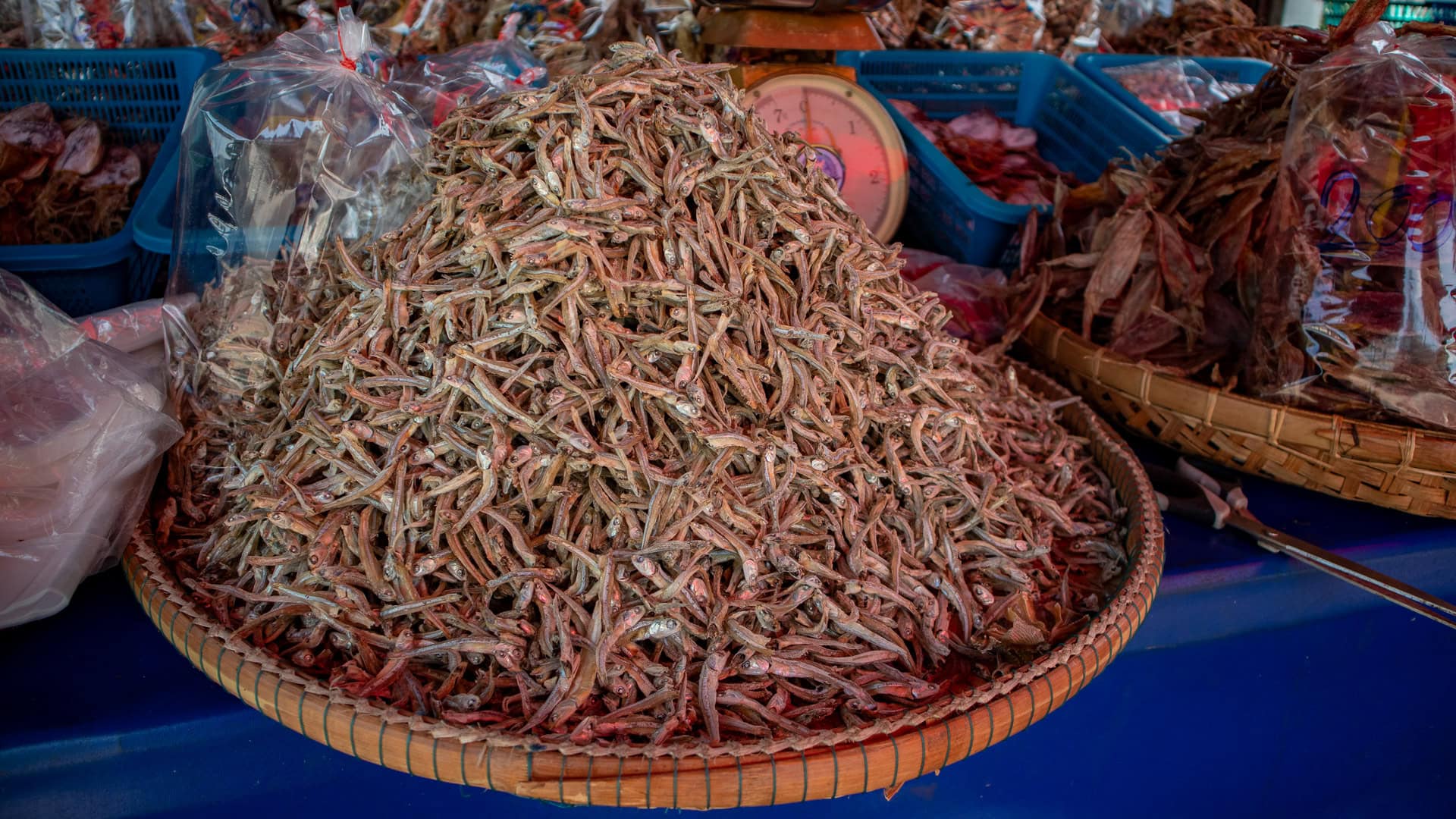 Large woven tray filled with small dried fish for sale, placed on a table among other dried seafood baskets.