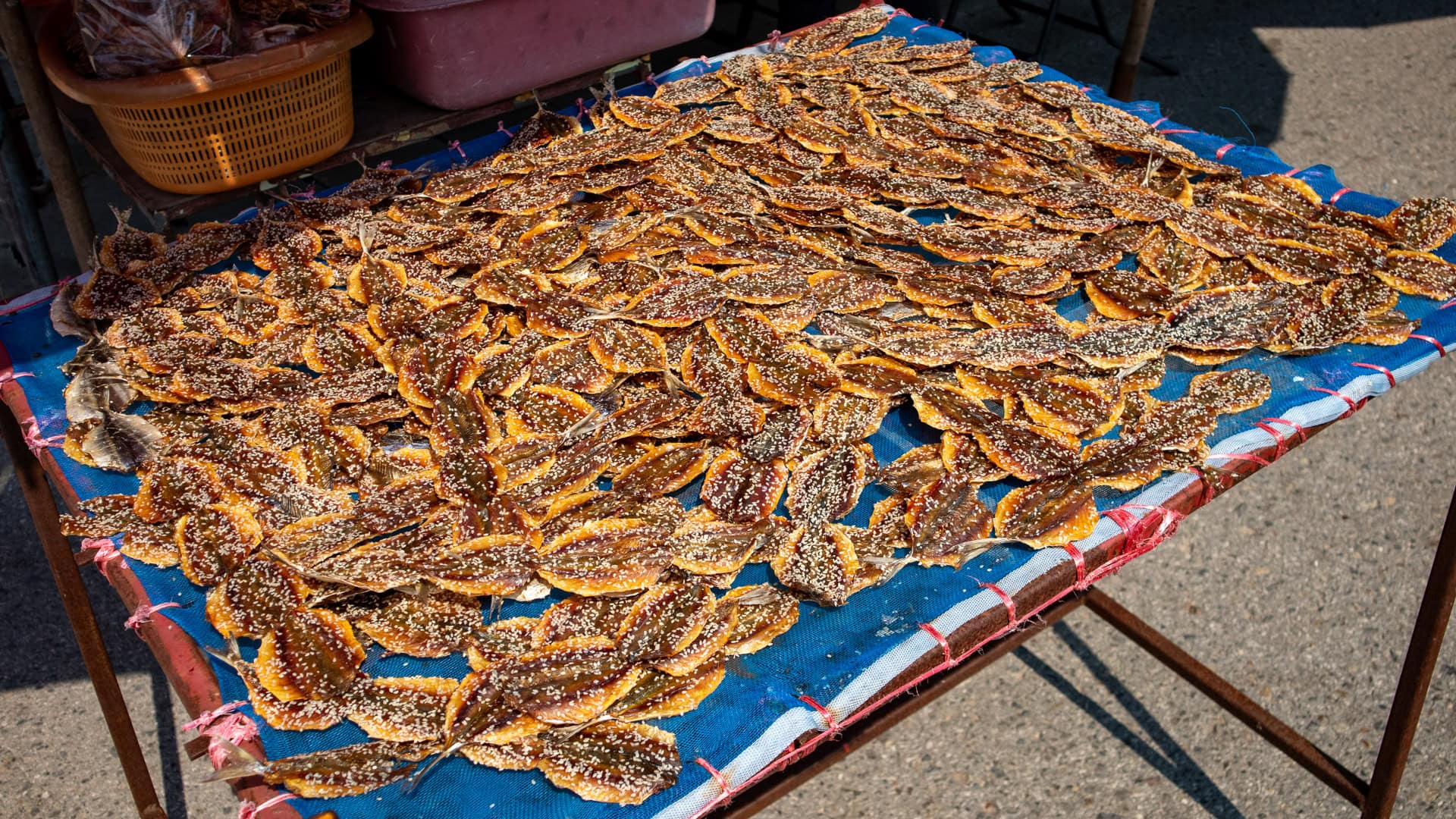Sweetened dried fish laid out on a mesh table outdoors, drying in direct sunlight beside the road.