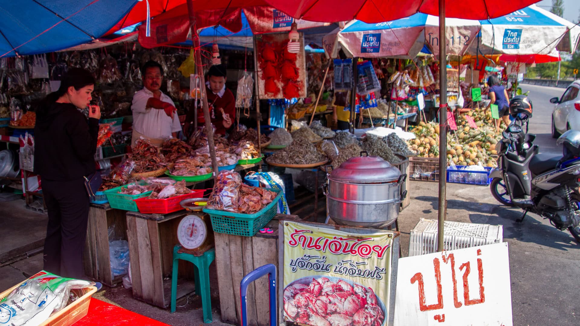 Busy roadside dried fish market stall with vendors, customers, scales, baskets of seafood, and umbrellas providing shade.