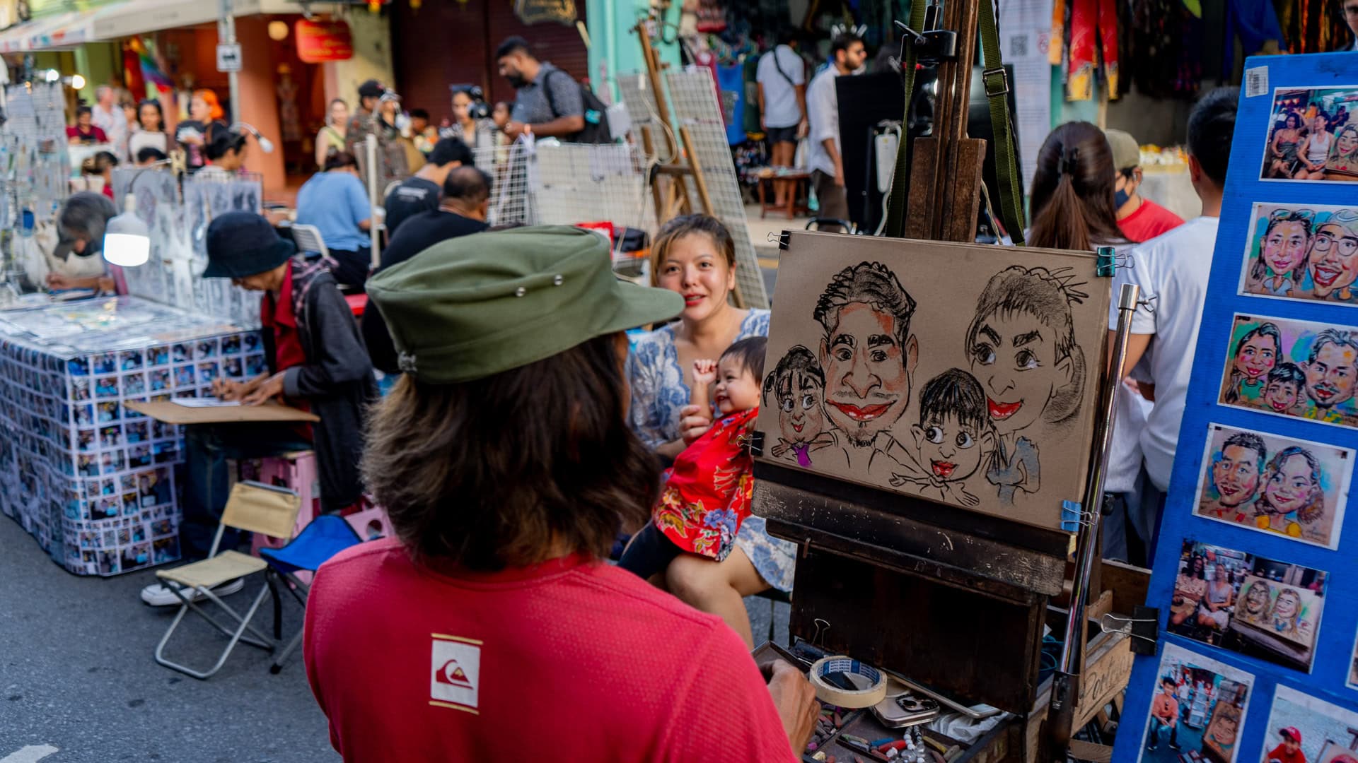 Lard Yai Sunday Night Market Phuket street caricature stall with an artist drawing a family portrait on a mounted board, a woman holding a child seated in front, and people gathered closely around the stand.