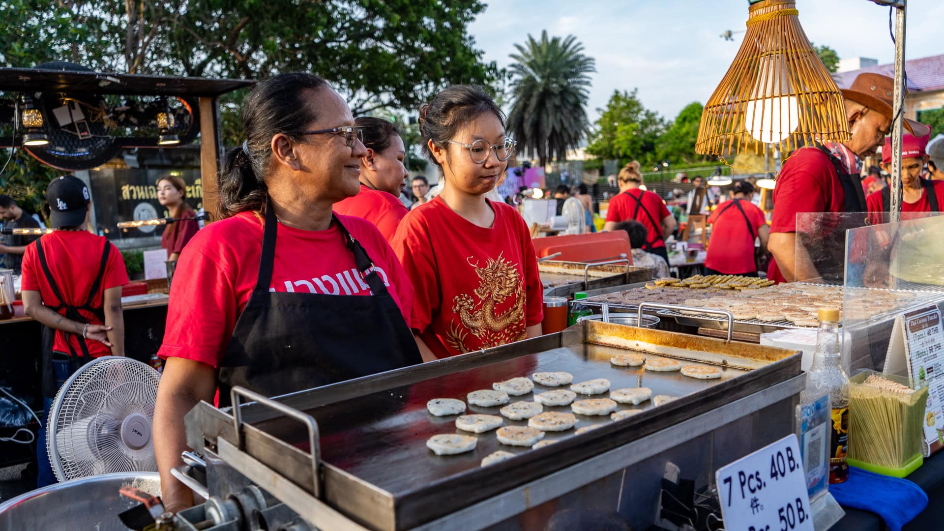 Lard Yai Sunday Night Market Phuket food stall with two women in red shirts standing behind a flat grill cooking small round cakes, rows of food arranged on the hot surface, and other vendors working at neighbouring stalls in the background.
