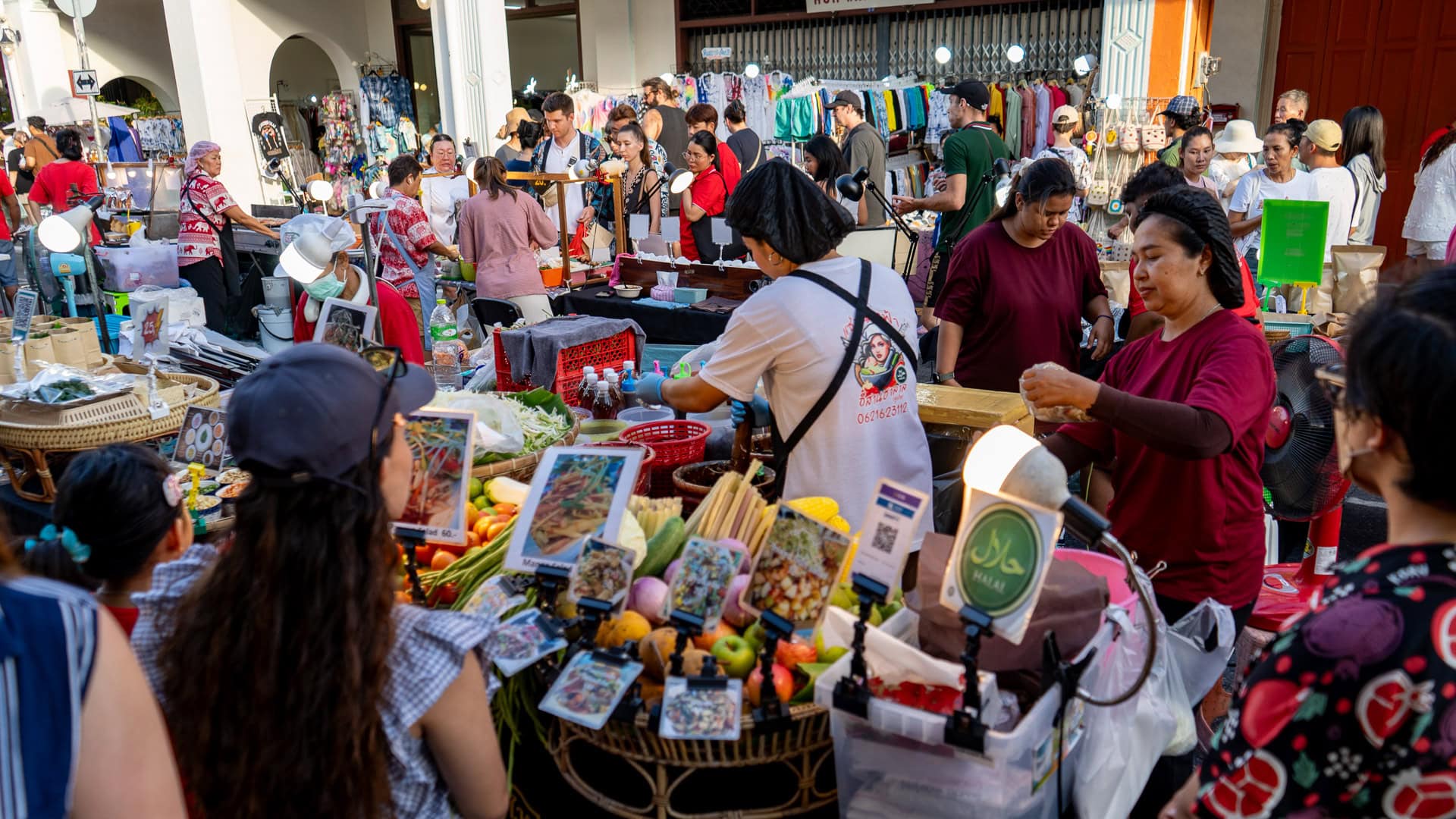 Lard Yai Sunday Night Market Phuket crowded food stall with several women preparing dishes from fresh vegetables and ingredients, customers gathered closely at the counter, and rows of clothing stalls visible behind the food area.
