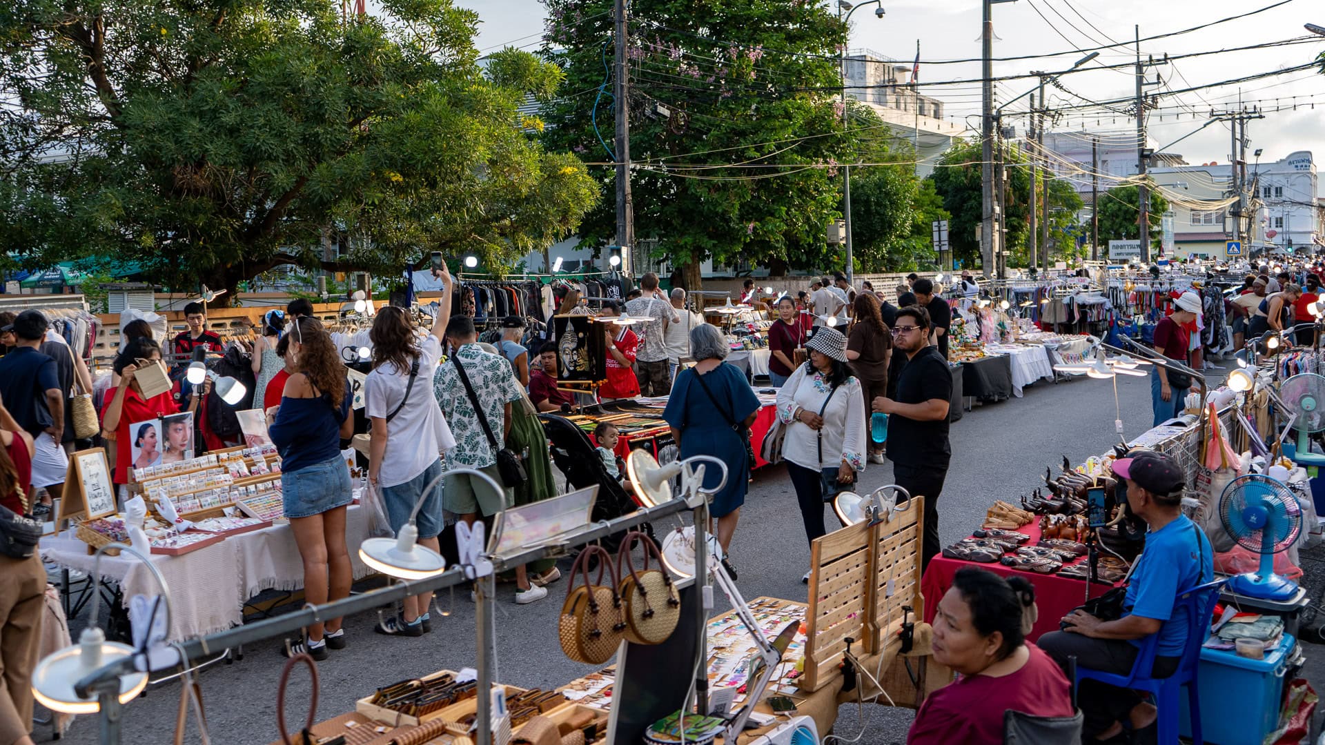 Lard Yai Sunday Night Market Phuket wide view of the market street with clothing and craft stalls set along the road, portable lights above tables, vendors seated behind displays, and visitors walking and browsing between the stalls.