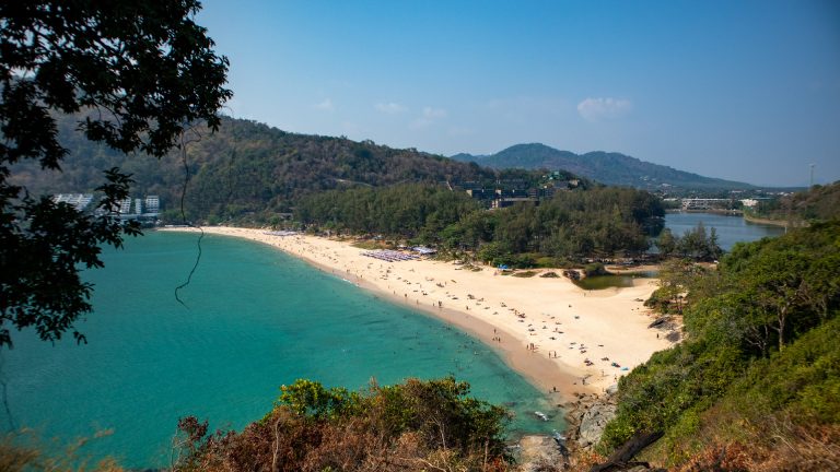Panoramic view of Nai Harn Beach Phuket backed by trees and hills.