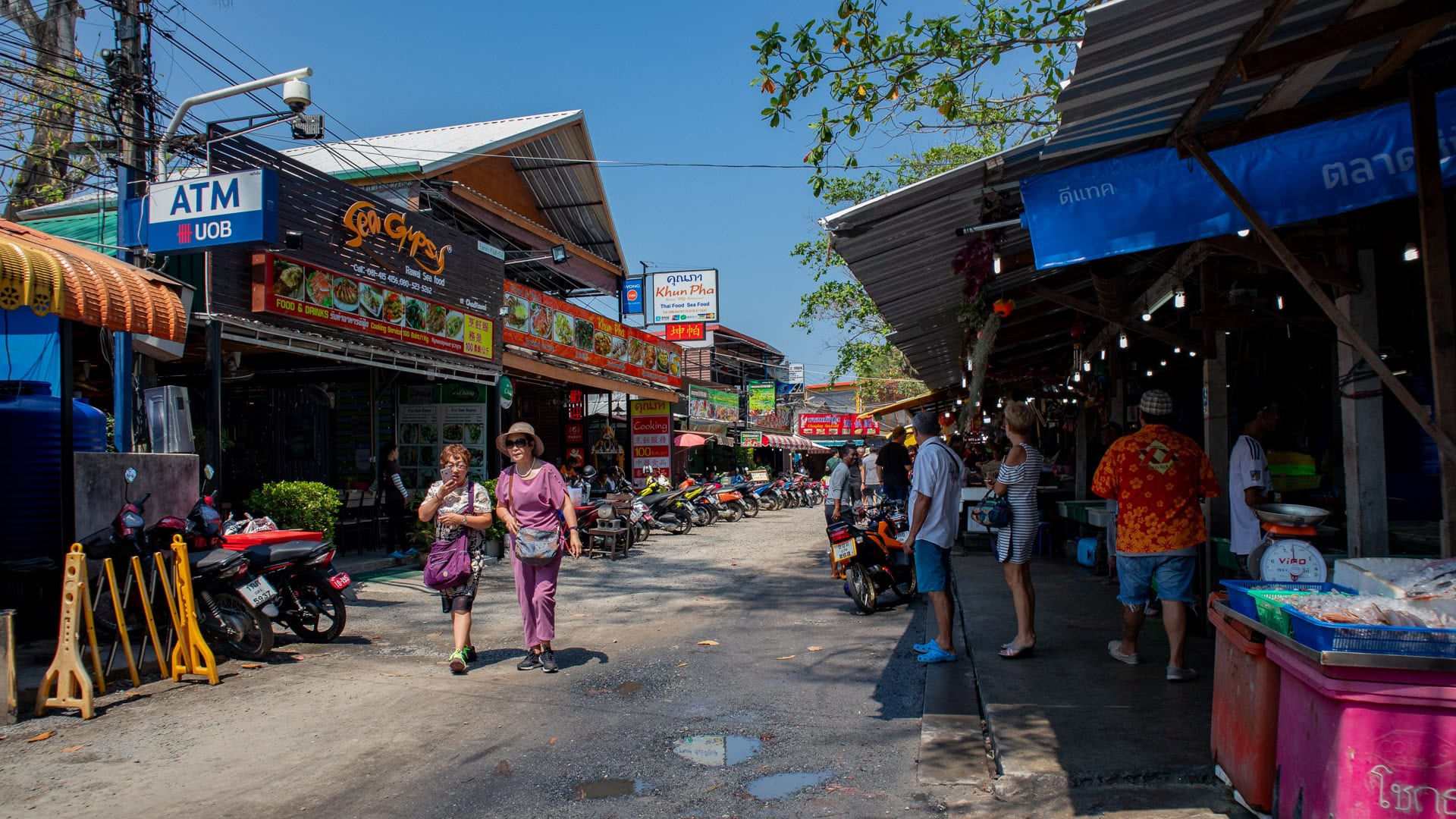 Street view of Rawai Seafood Market with seafood stalls on one side, restaurants on the other, and people walking through the open road between them.