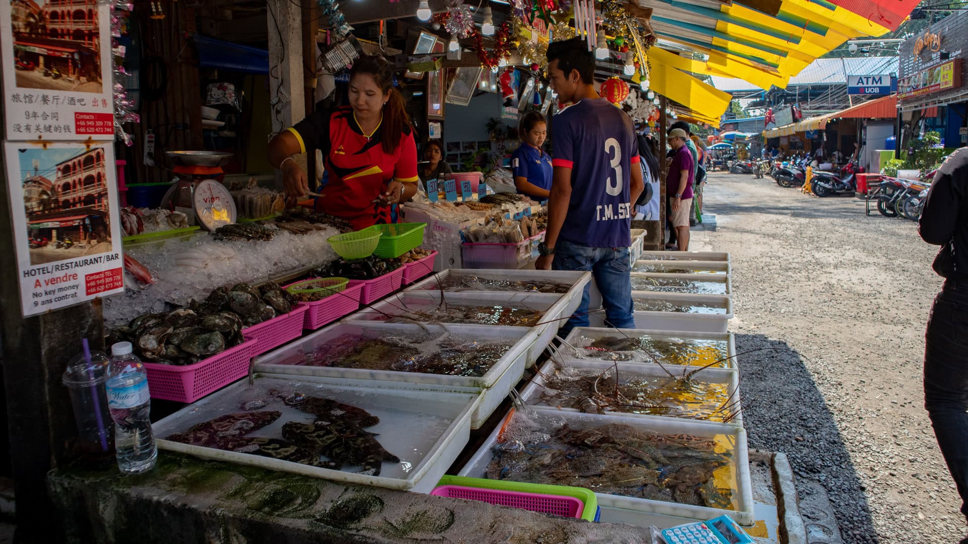 Live seafood tanks and trays along the roadside at Rawai Seafood Market with vendors and customers selecting fish and shellfish.