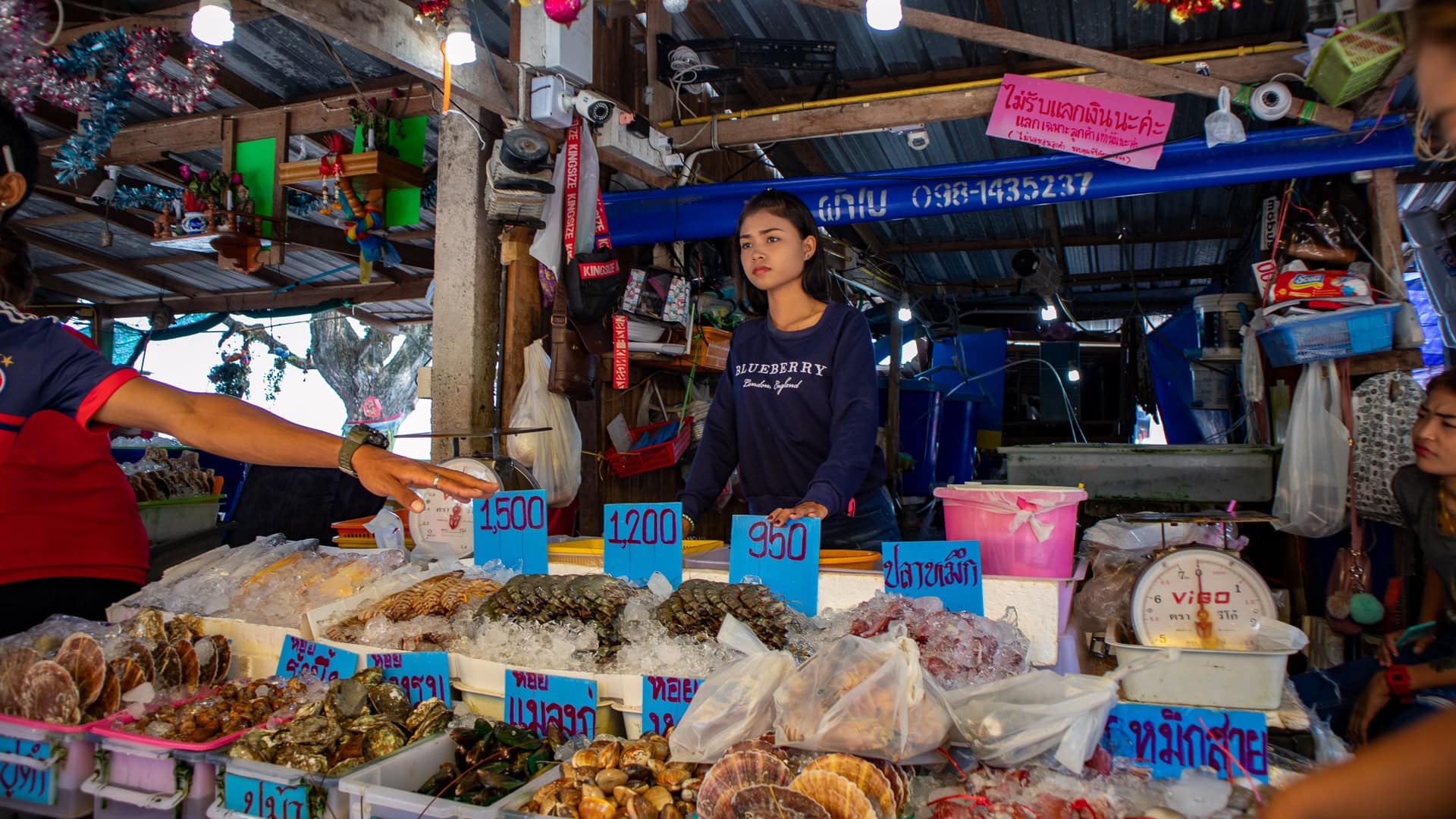 Seafood vendor weighing and pricing shellfish and fish on ice at Rawai Seafood Market, with prices displayed in Thai baht.