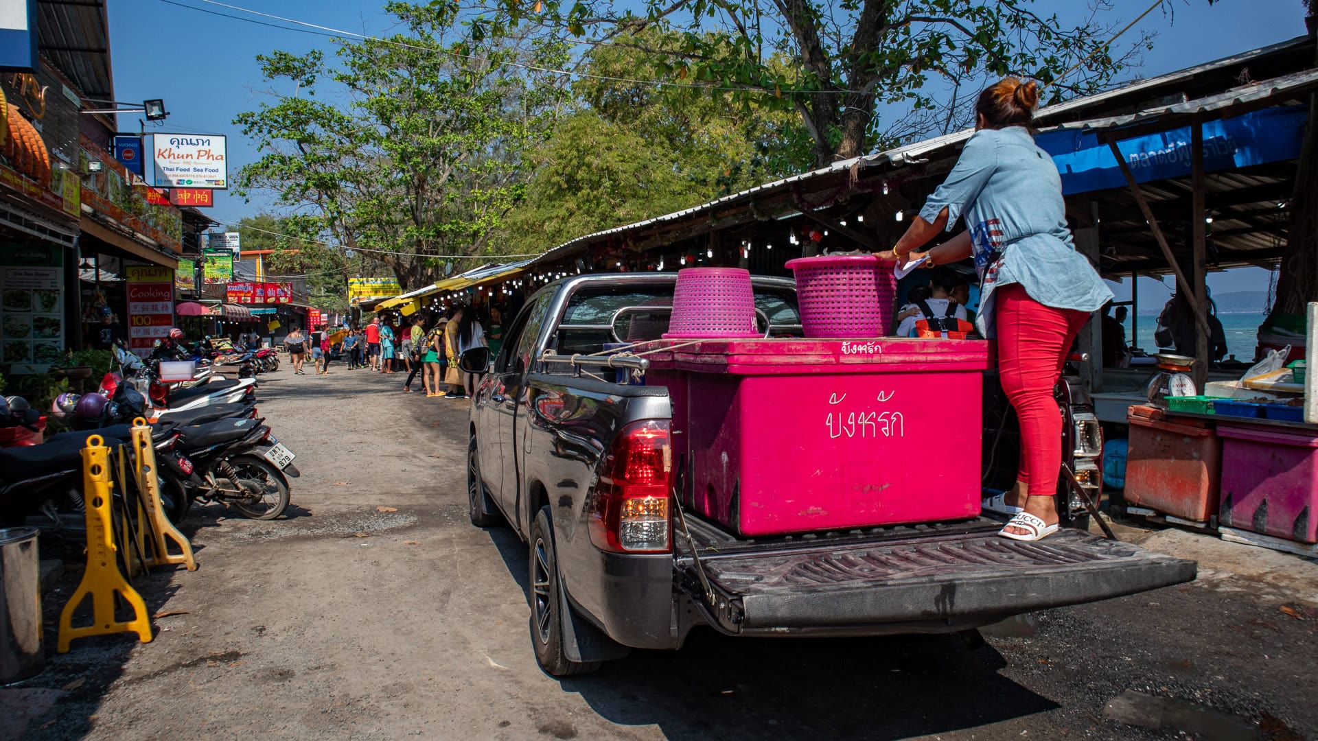 Pickup truck unloading large insulated containers in the middle of Rawai Seafood Market during active trading hours.