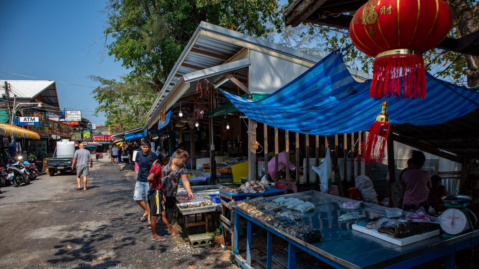 Seafood tables set along the edge of Rawai Seafood Market near the water, with people resting in the shade behind the stalls.