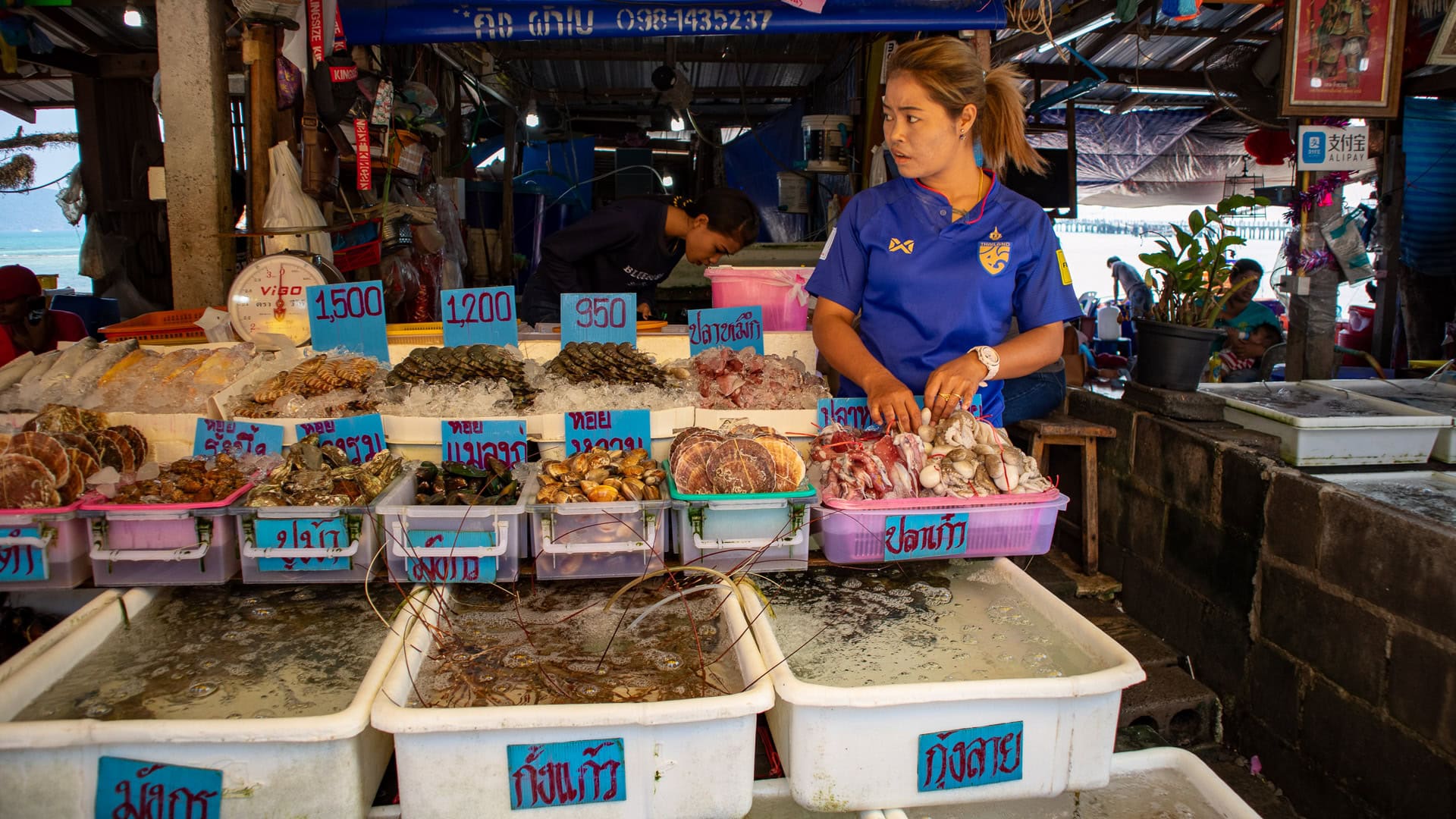 Seafood vendor standing behind ice-filled trays at Rawai Seafood Market, briefly looking away from her work toward something off-frame.