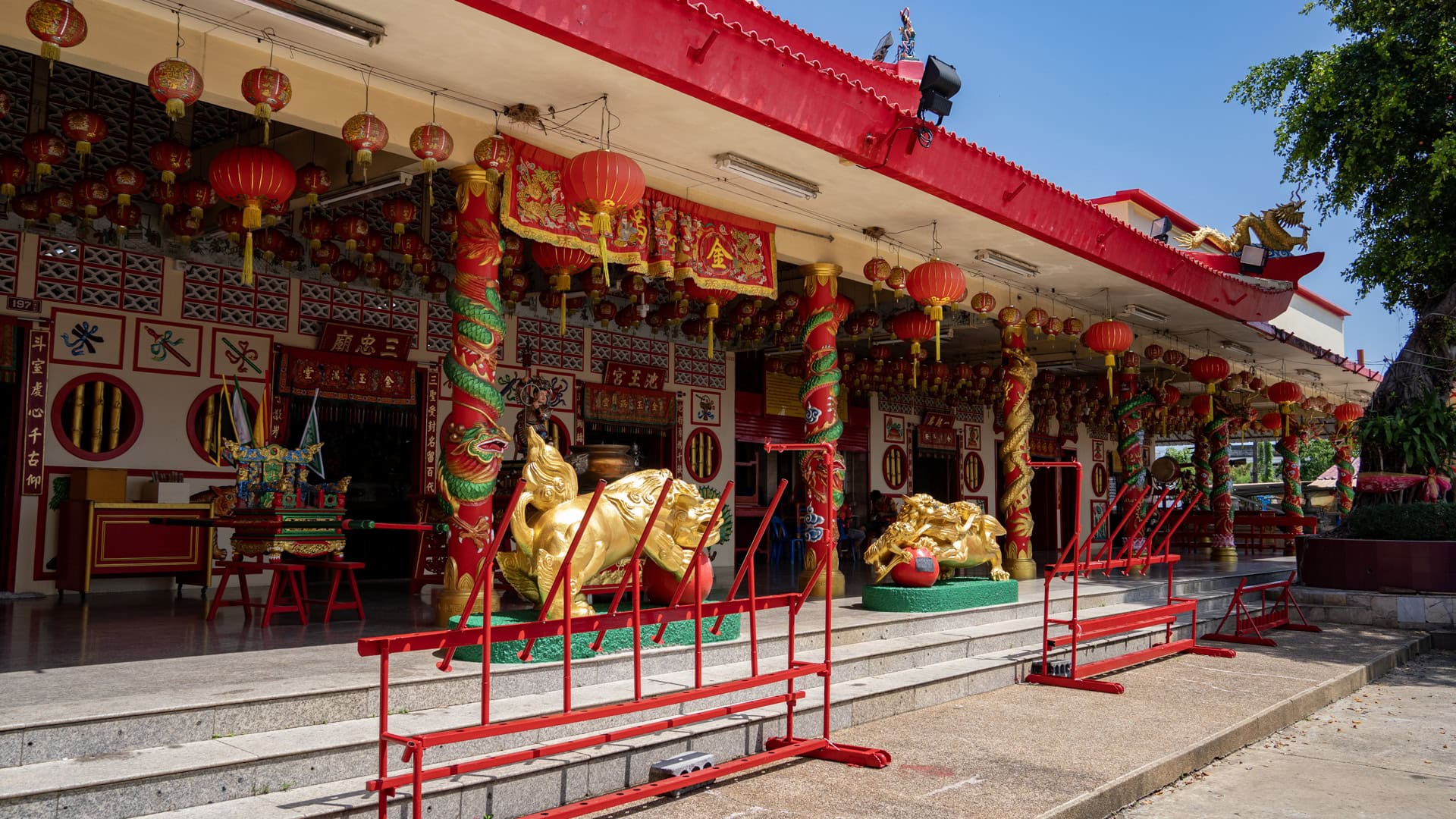 Angled view of the entrance porch at Bang Neow Shrine with red lanterns, dragon-wrapped columns, gold guardian statues, and red railings.