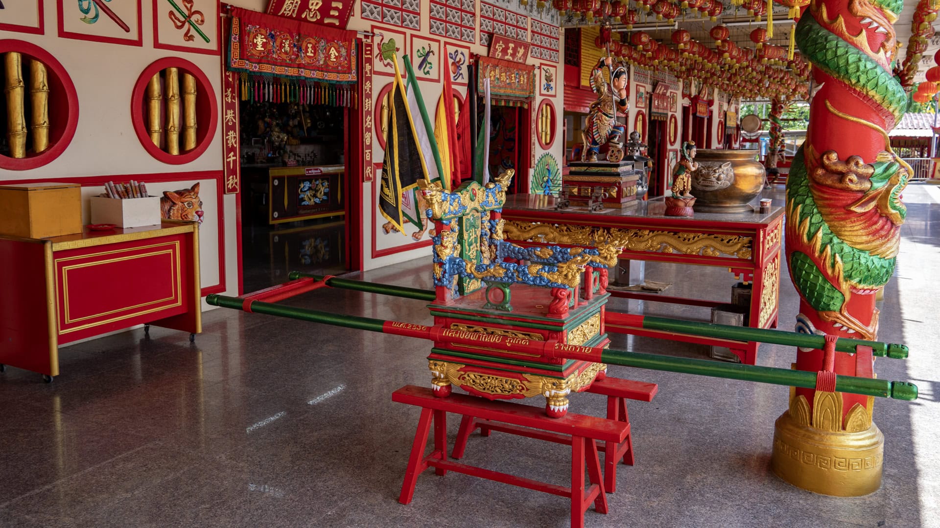 A painted portable shrine sedan with long carrying poles stands in front of an altar table and dragon-wrapped column inside Bang Neow Shrine.
