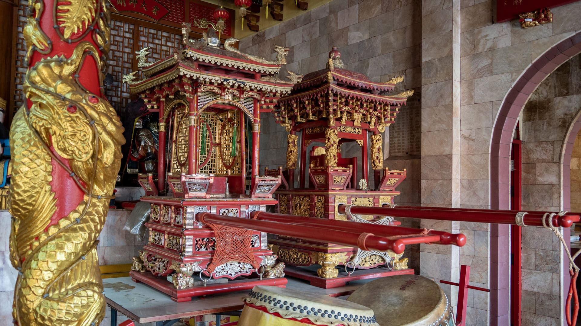 Two ornate red-and-gold sedan chairs with long poles stand beside a drum inside a stone-walled chamber at Bang Neow Shrine.Two ornate red-and-gold sedan chairs with long poles stand beside a drum inside a stone-walled chamber at Bang Neow Shrine.