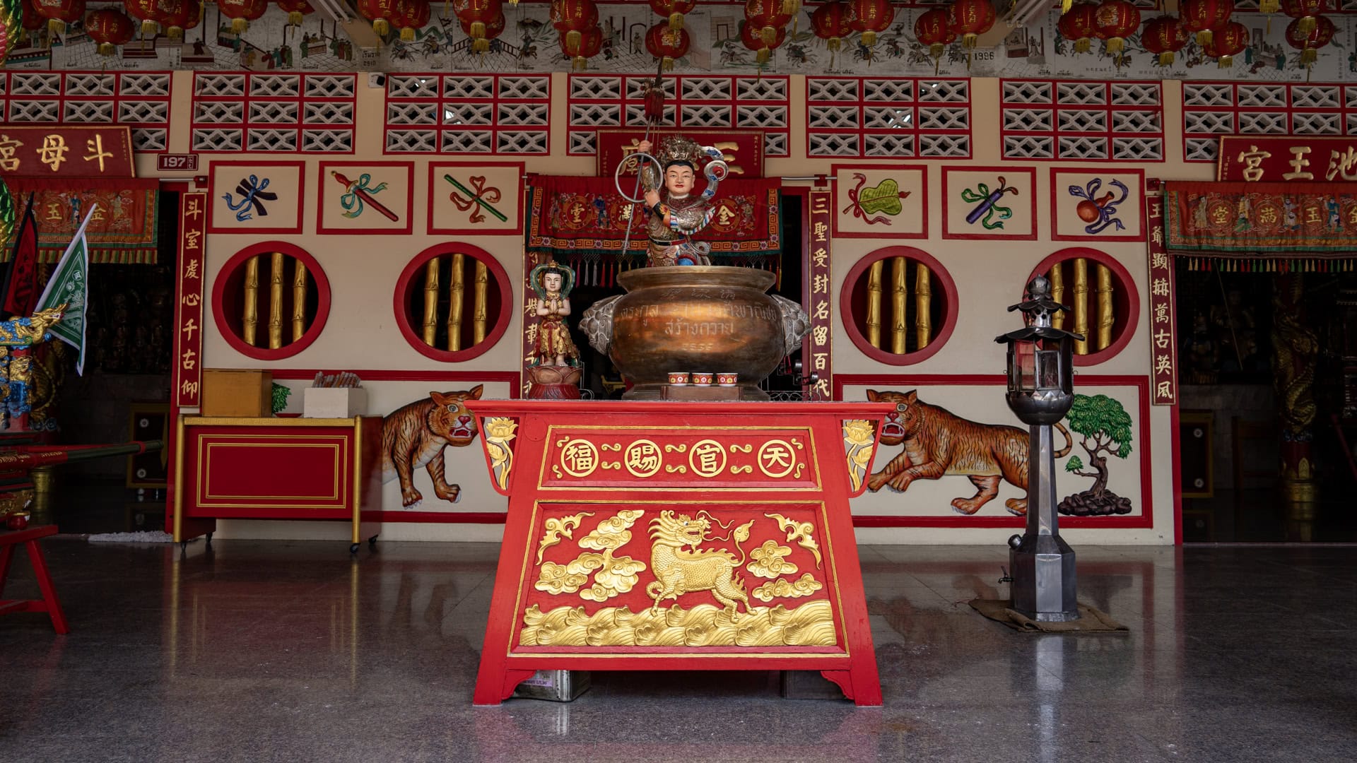 Bronze incense burner on a red altar stand in front of the shrine wall, with a small figure above and tiger paintings on either side.