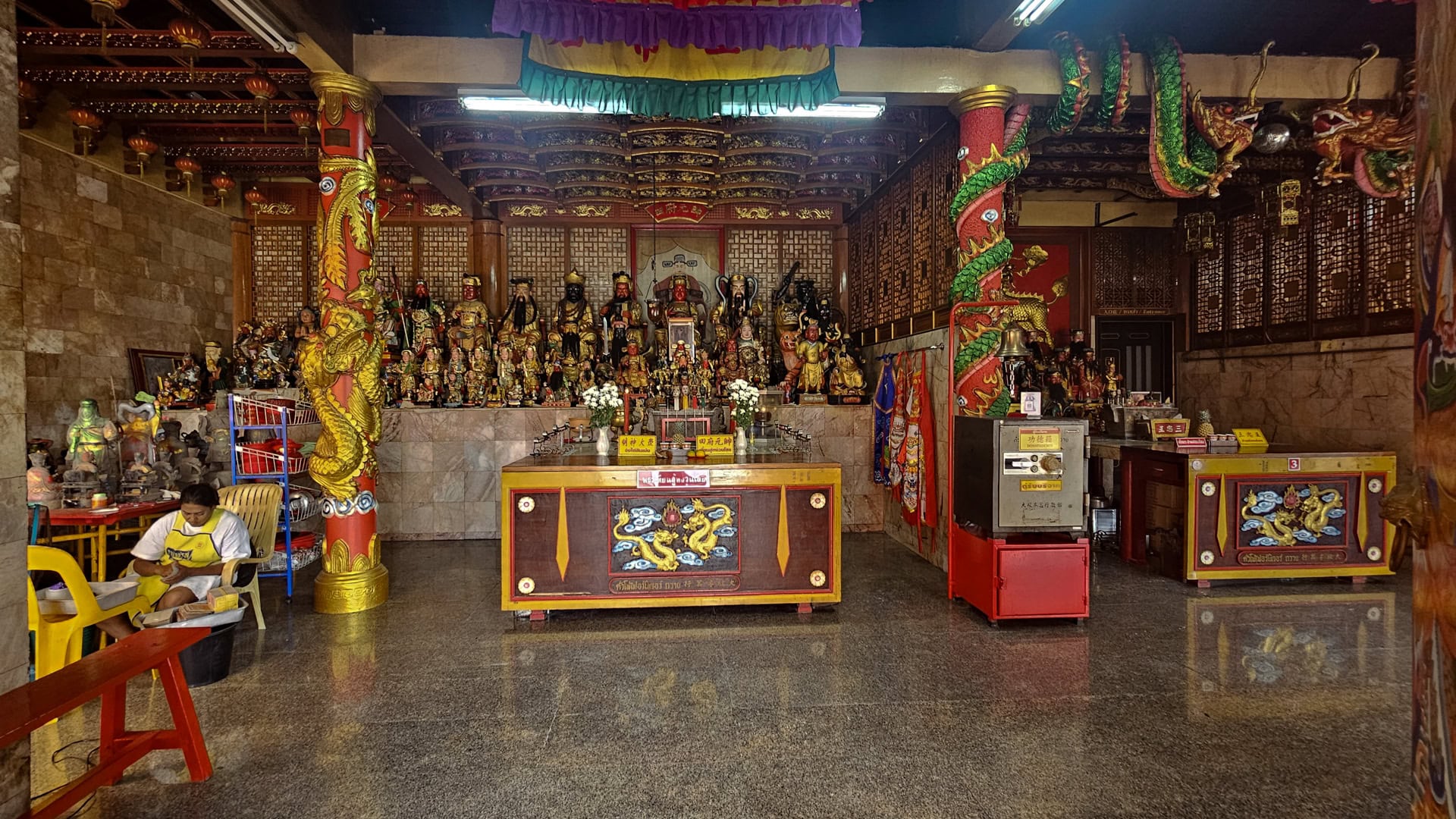 Wide interior view of Bang Neow Shrine showing dragon-wrapped columns, altar tables, many devotional figures, and a seated man at left.
