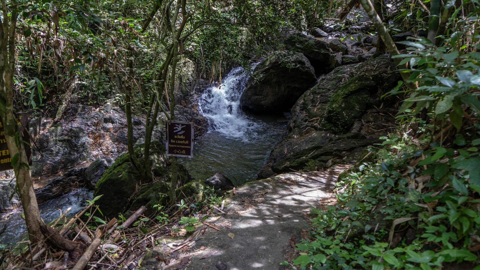 A small cascade at Bang Pae Waterfall drops into a narrow pool beside a concrete path, with a warning sign reading “Be careful” hanging among the trees.