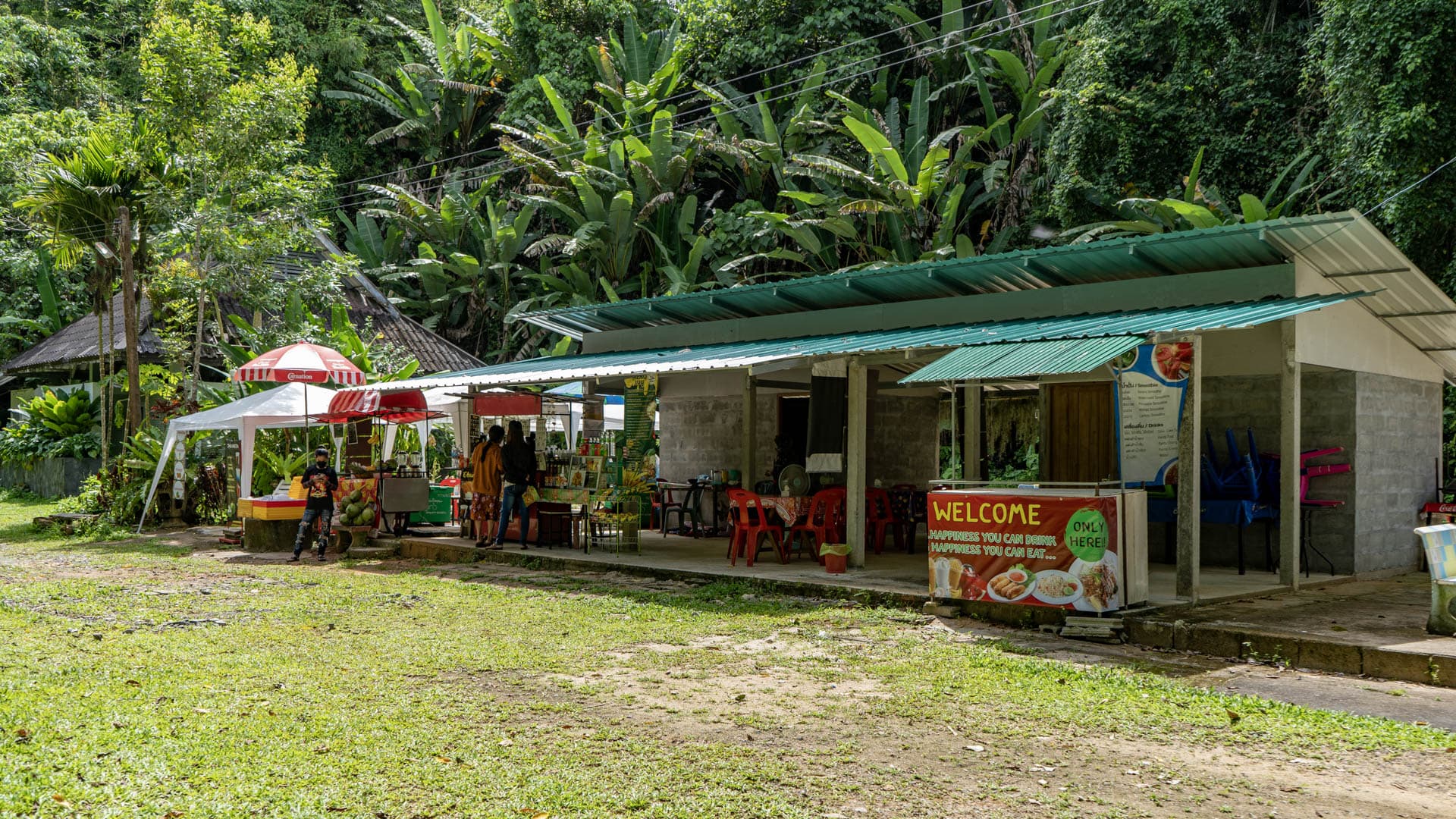 Refreshment stalls and an open pavilion stand near Bang Pae Waterfall, with red umbrellas, plastic chairs, and jungle vegetation rising behind the buildings.