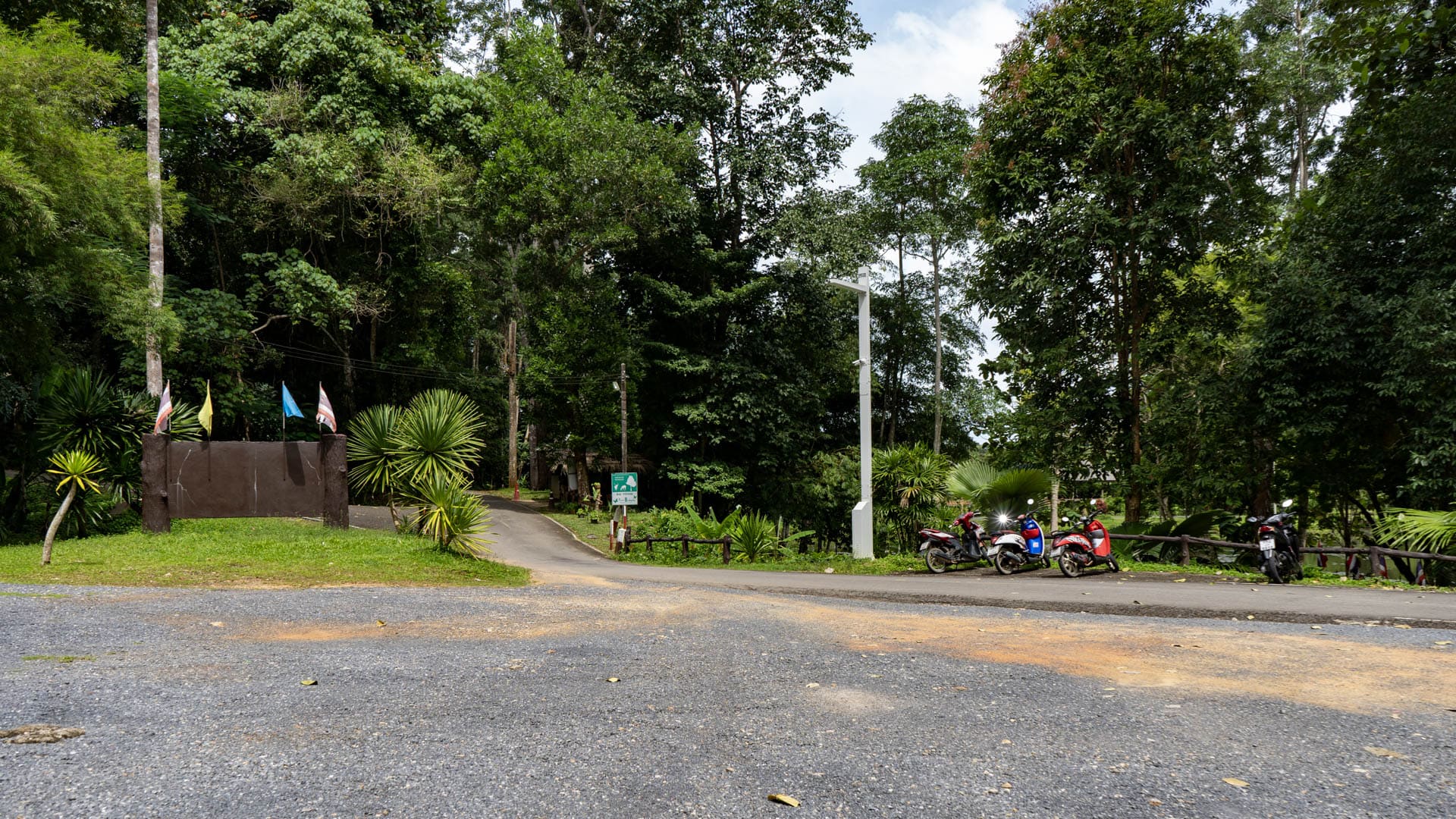 The entrance road at Bang Pae Waterfall curves into dense trees, with parked motorbikes, small flags, and roadside planting along the edge.