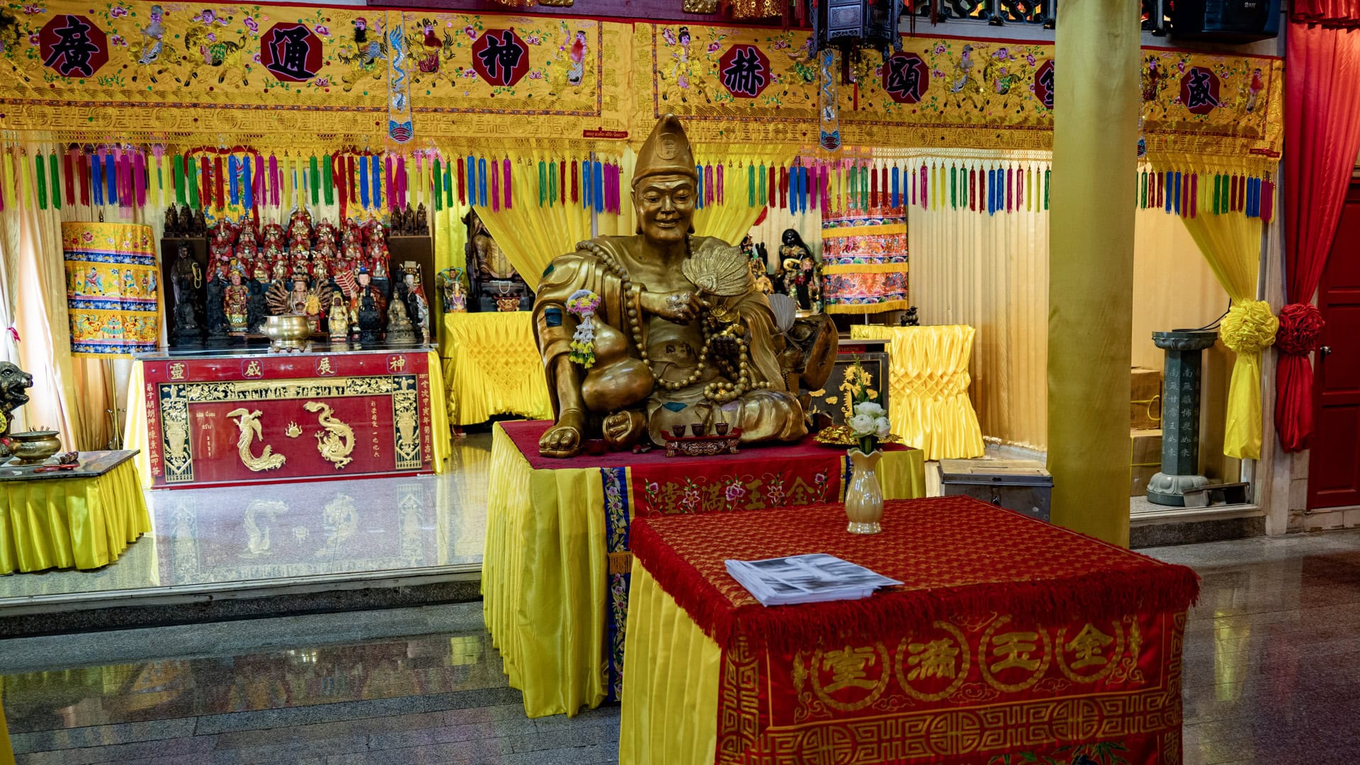 A seated gold-toned deity figure at San Chao Jui Tui sits on a red-and-yellow table in front of a brightly decorated altar with tassels, figures, and yellow drapery.