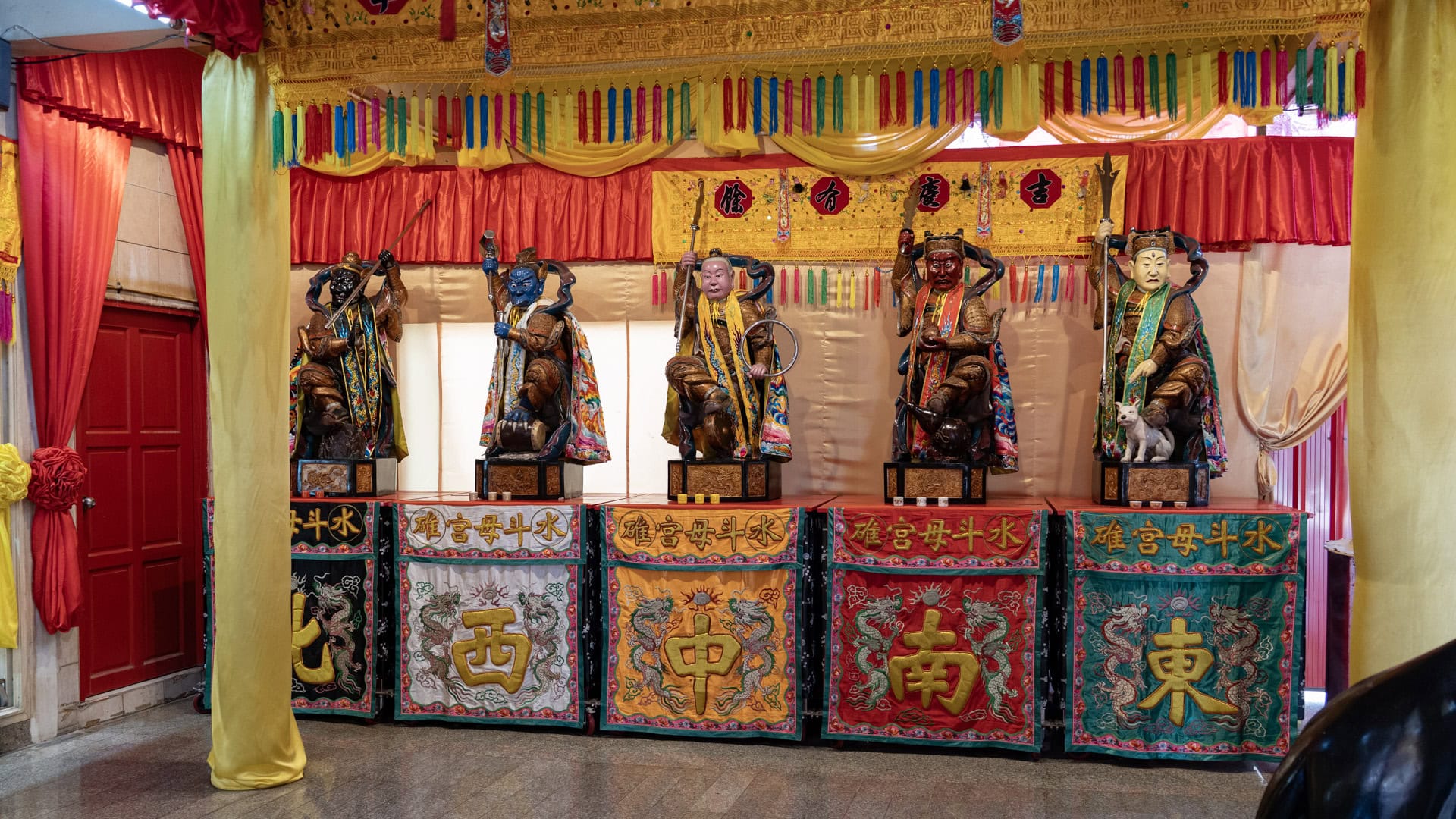 Five painted guardian figures at San Chao Jui Tui sit on raised platforms inside the shrine, each backed by drapery and fronted by embroidered cloth panels.