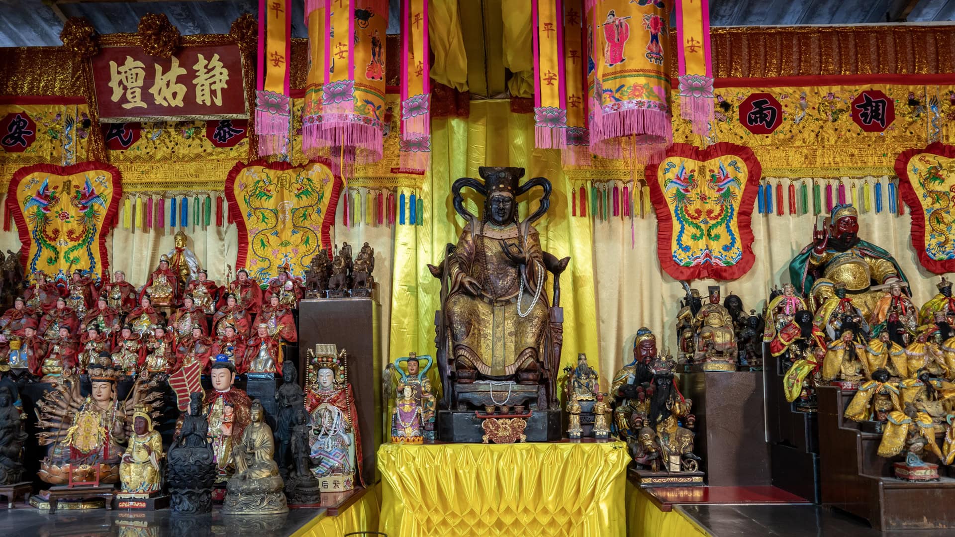 A central seated figure at San Chao Jui Tui stands on a yellow-covered altar table, surrounded by many smaller shrine figures and tall hanging banners in pink and yellow.