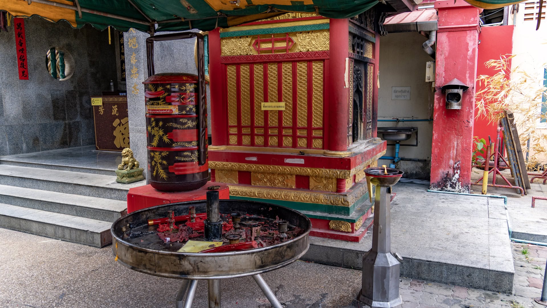 A red-and-gold firecracker tower at San Chao Jui Tui stands beside a round metal burner filled with red wax and debris, with a small flame burning on a stand nearby.