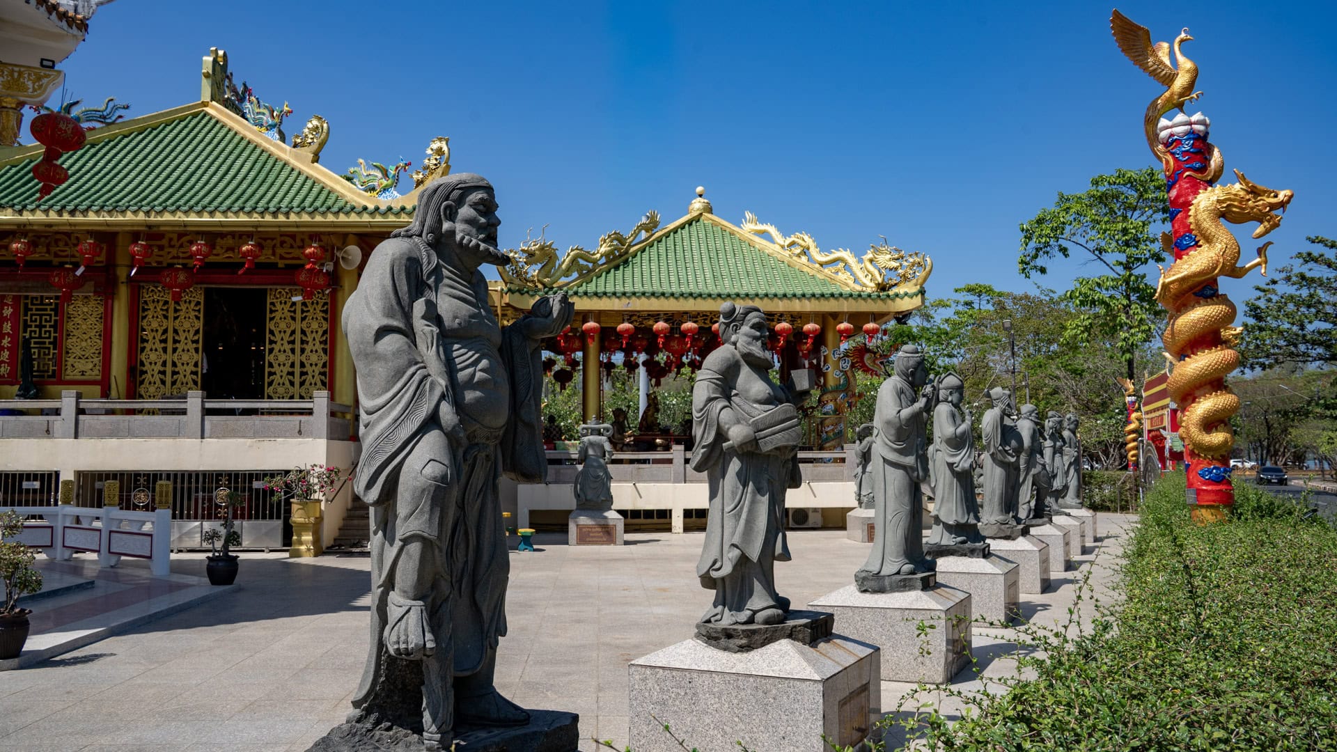 Stone figures stand in a row across the forecourt at San Chao Kiew Tien Keng, with green-tiled shrine roofs and a gold dragon-wrapped column behind them.