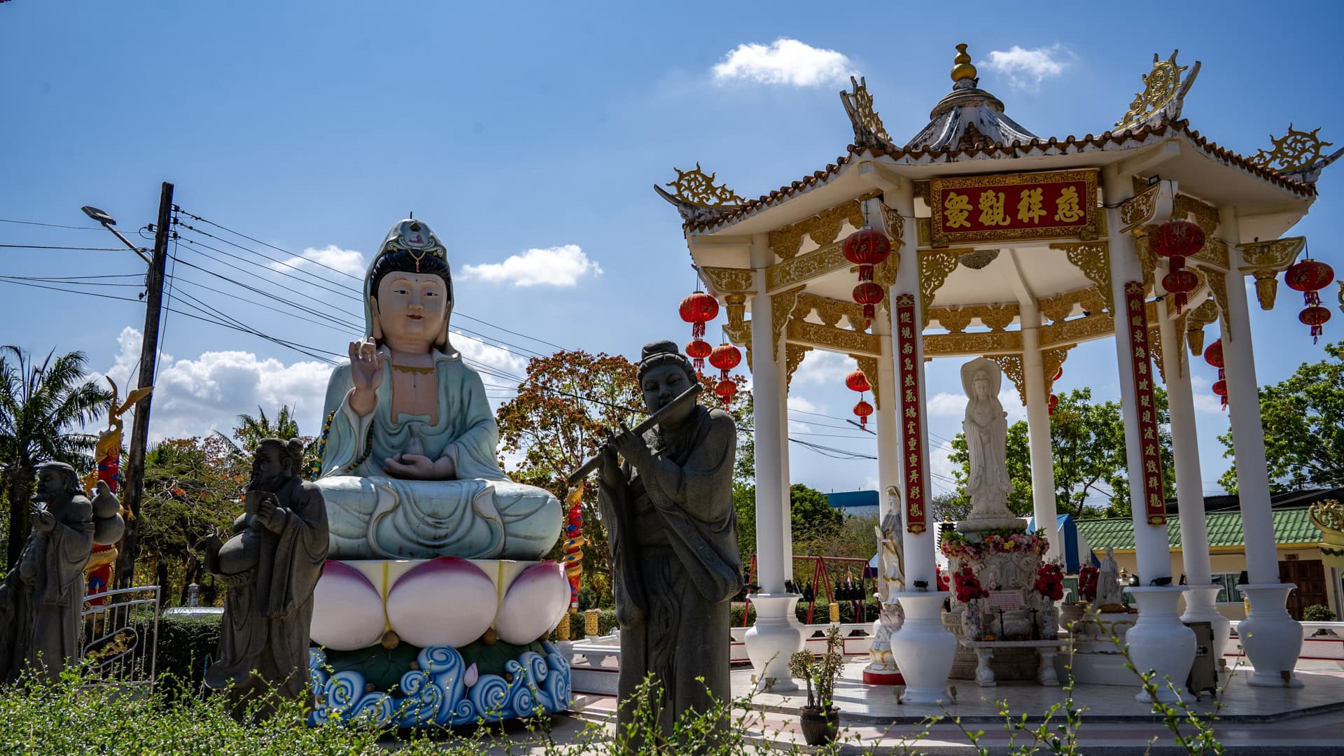 A seated Guanyin statue at San Chao Kiew Tien Keng stands beside a white-and-gold pavilion hung with red lanterns, with smaller stone figures in the foreground.