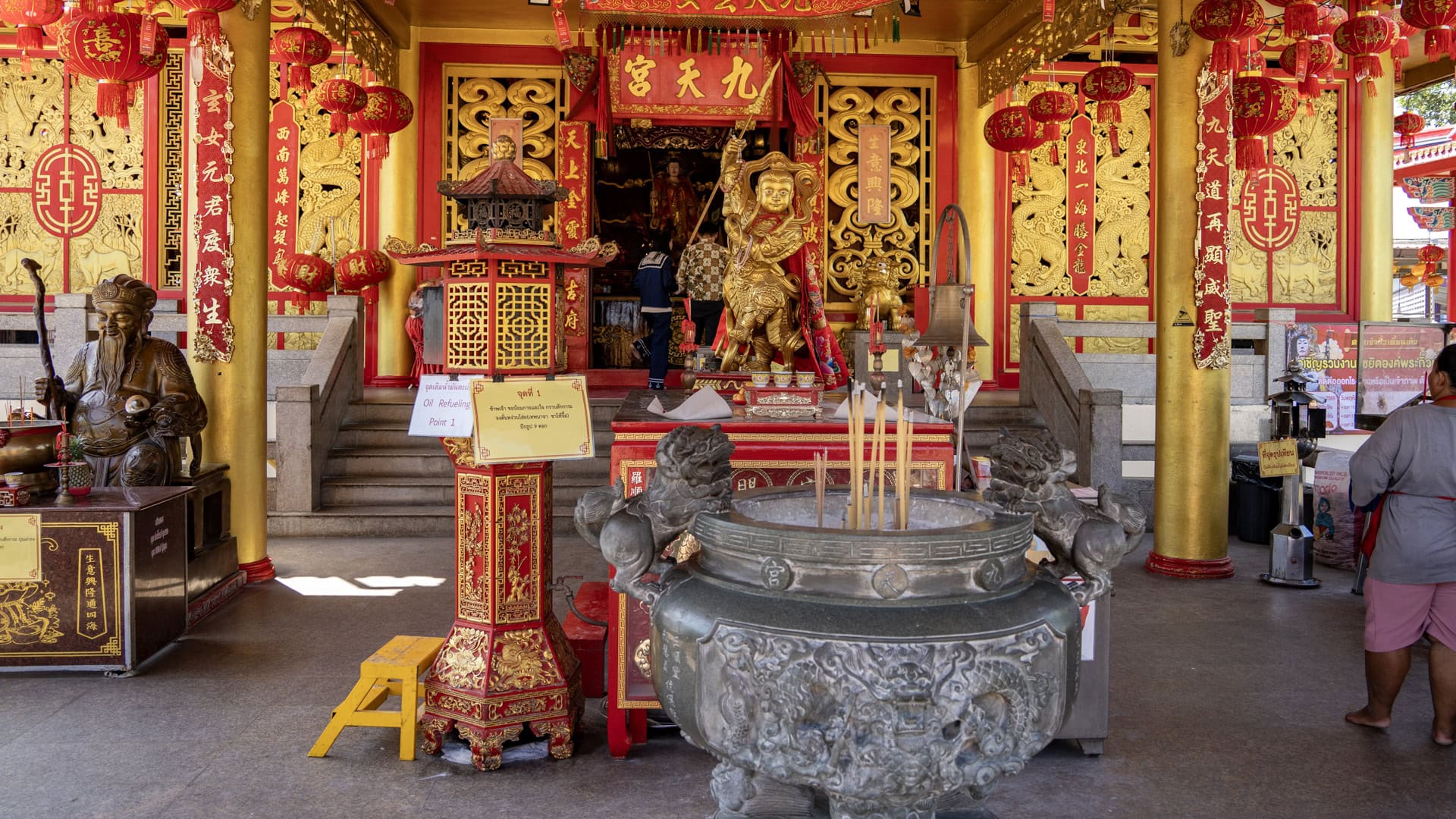 A large metal incense urn stands before the main hall at San Chao Kiew Tien Keng, with red lanterns, gold panels, and a central gilded figure beyond.