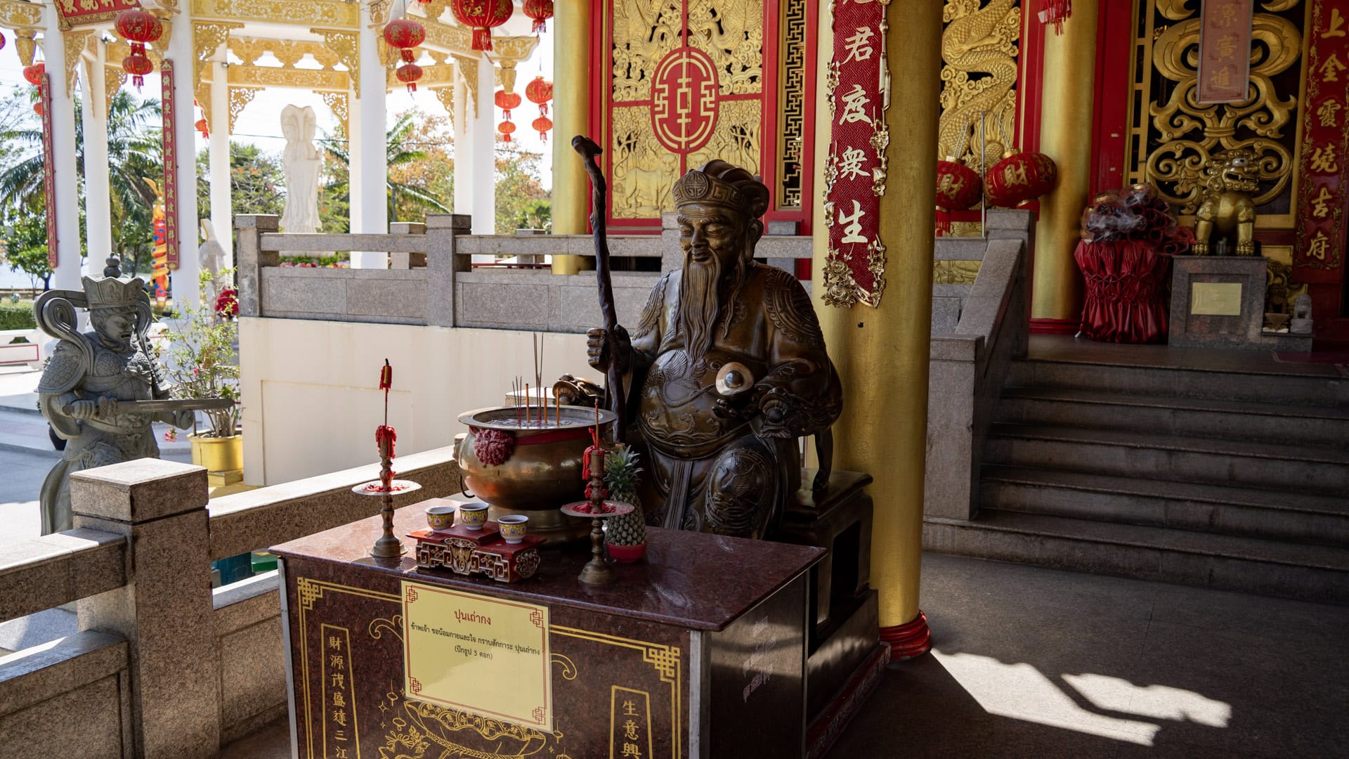 A seated bronze deity statue at San Chao Kiew Tien Keng stands beside a small offering table with incense, with the hall steps and pavilion beyond.