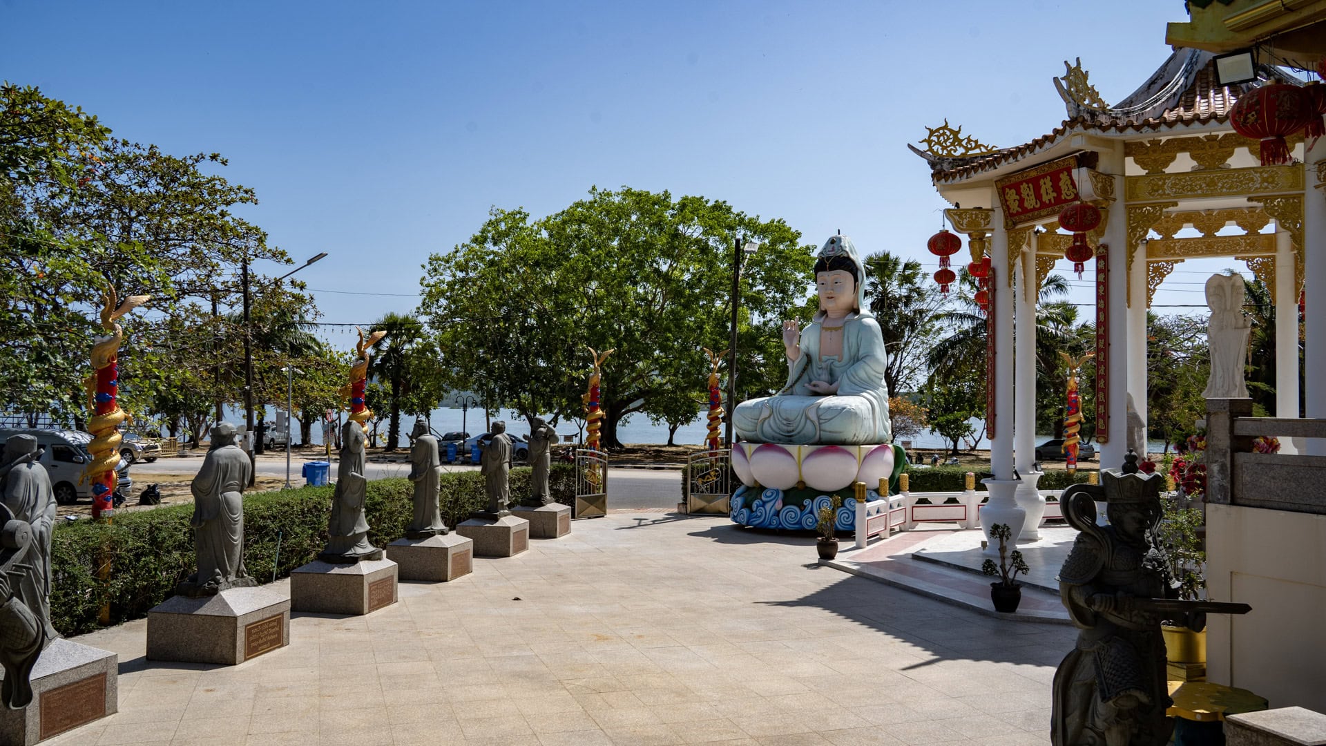 A seated Guanyin statue at San Chao Kiew Tien Keng faces an open forecourt lined with stone figures, with trees and the sea visible beyond the roadside edge.