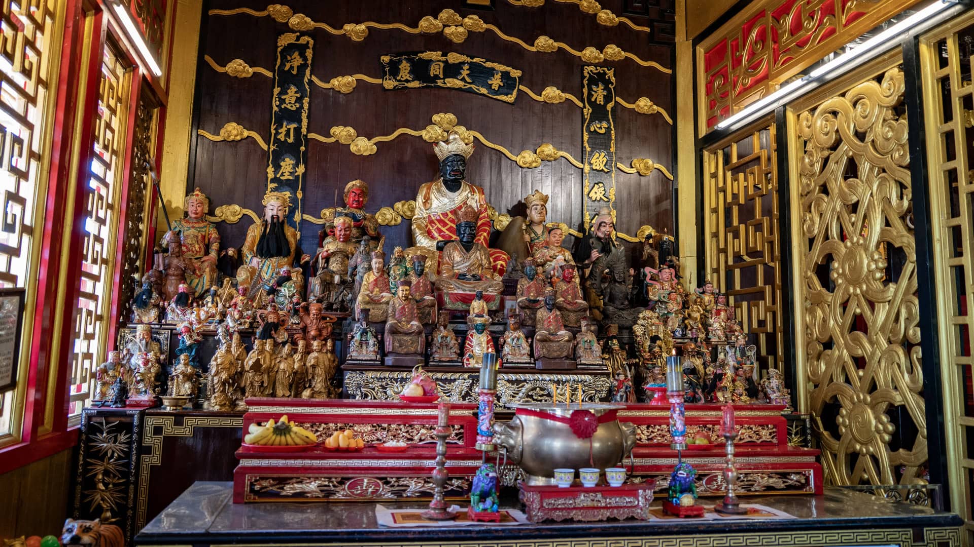 A side altar at San Chao Kiew Tien Keng holds many deity figures of different sizes, with a black-faced central figure, incense burner, and gold carved panels around the niche.