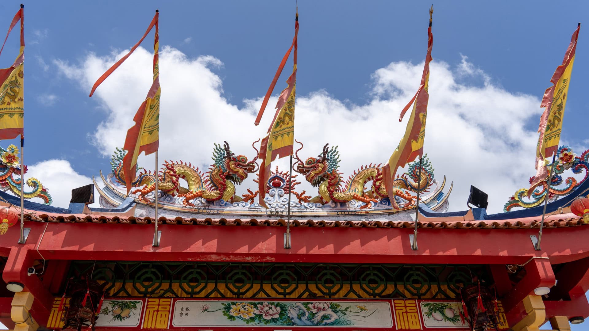 Roof ridge at Sam Kong Shrine with two facing dragons and tall yellow-and-red flags rising in front of the eaves.