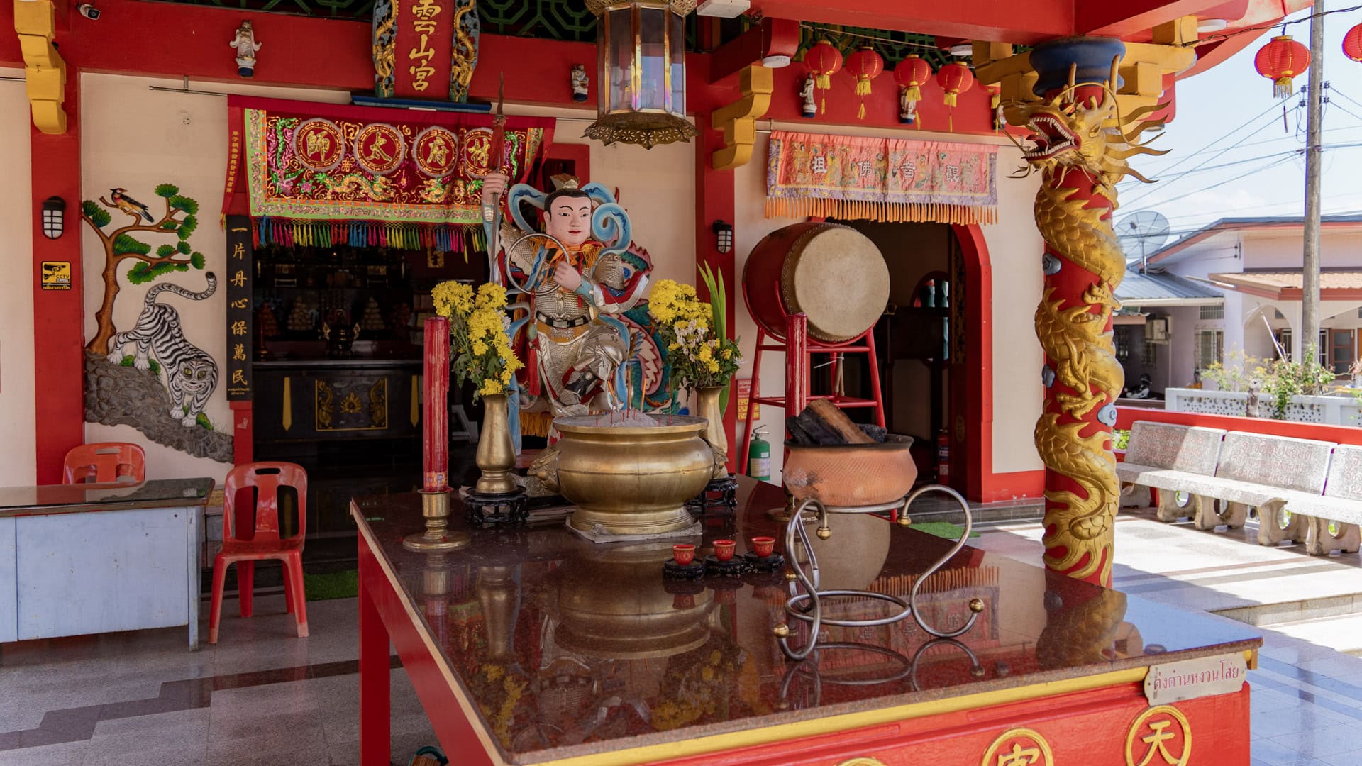 Front prayer area at Sam Kong Shrine with a brass incense burner, red candles, a guardian figure, a drum, yellow flowers, and a dragon-wrapped column.