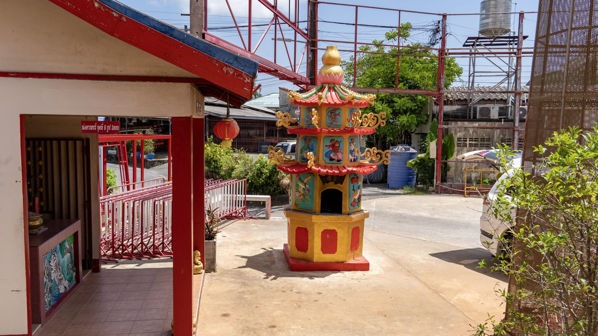 Small painted spirit tower in a side yard at Sam Kong Shrine, surrounded by concrete paving, red railings, utility frames, and nearby buildings.