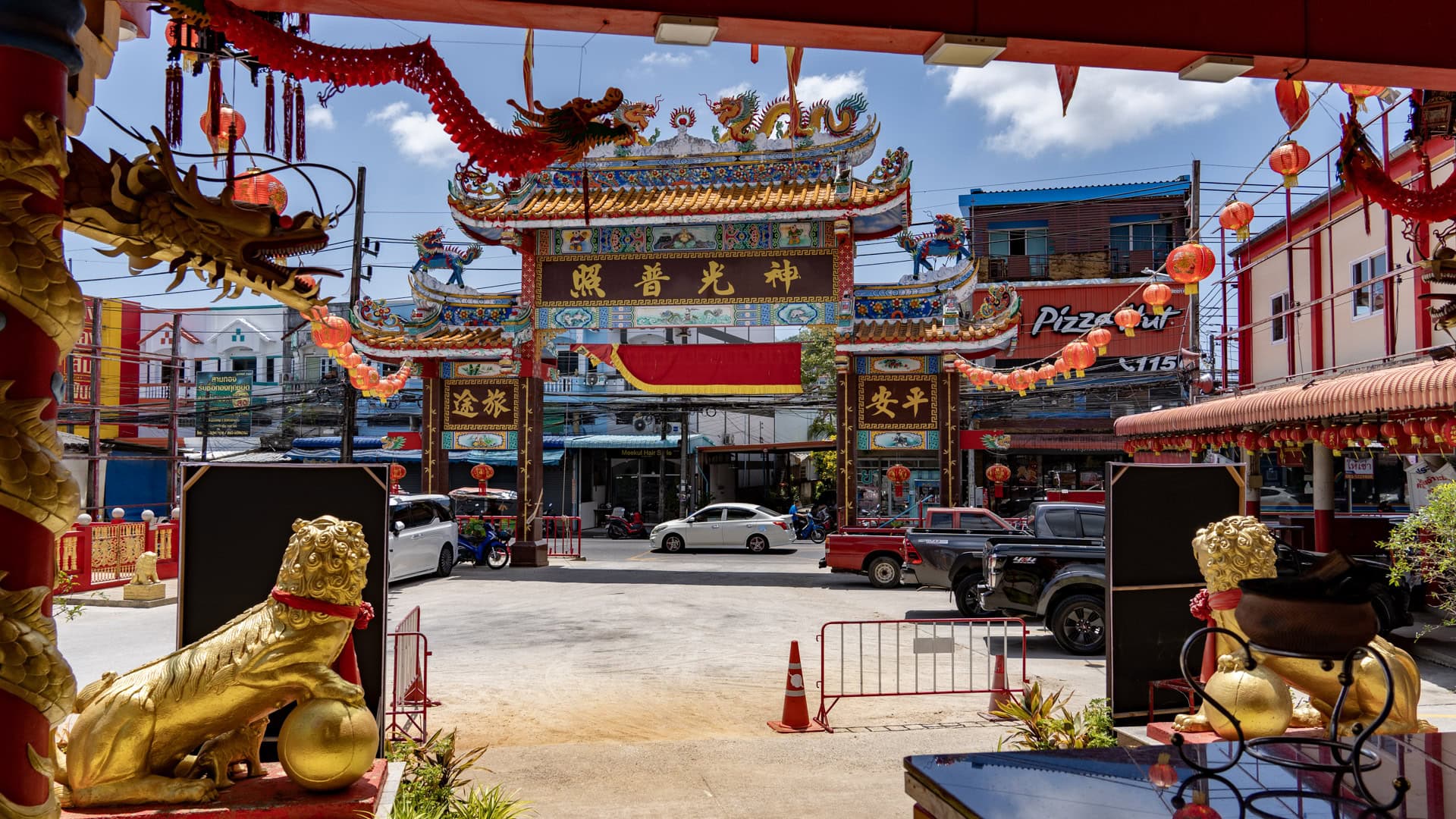 View from inside Sam Kong Shrine toward the entrance arch, framed by lanterns, a gold lion figure, a dragon head, and parked vehicles outside.