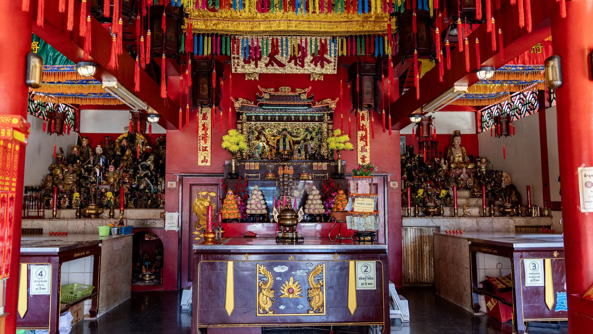 Main inner hall at Sam Kong Shrine with a central altar, hanging red tassels, side deity platforms, offering tables, and fruit offerings.