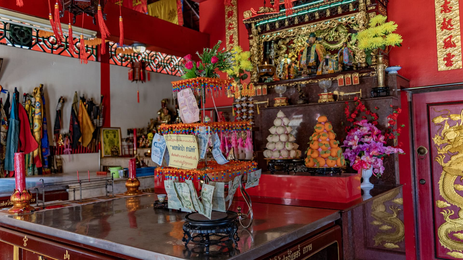 Decorated offering stand with hanging banknotes on a table inside Sam Kong Shrine, with fruit offerings, flowers, and the main altar behind it.