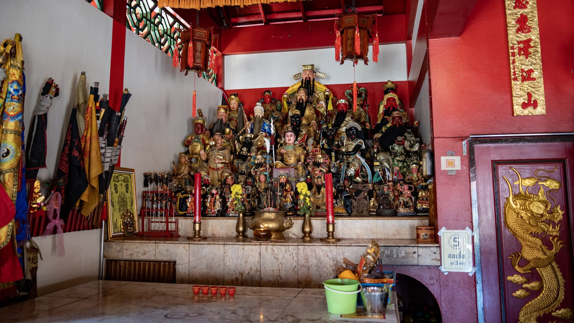 Side altar inside Sam Kong Shrine filled with many deity figures, red candles, hanging lanterns, yellow flowers, and ritual flags along the left wall.