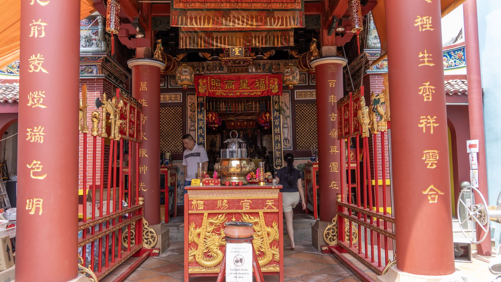 Red pillars frame the front hall at San Chao Saengtham, with a brass incense burner at the centre and two people visible inside.