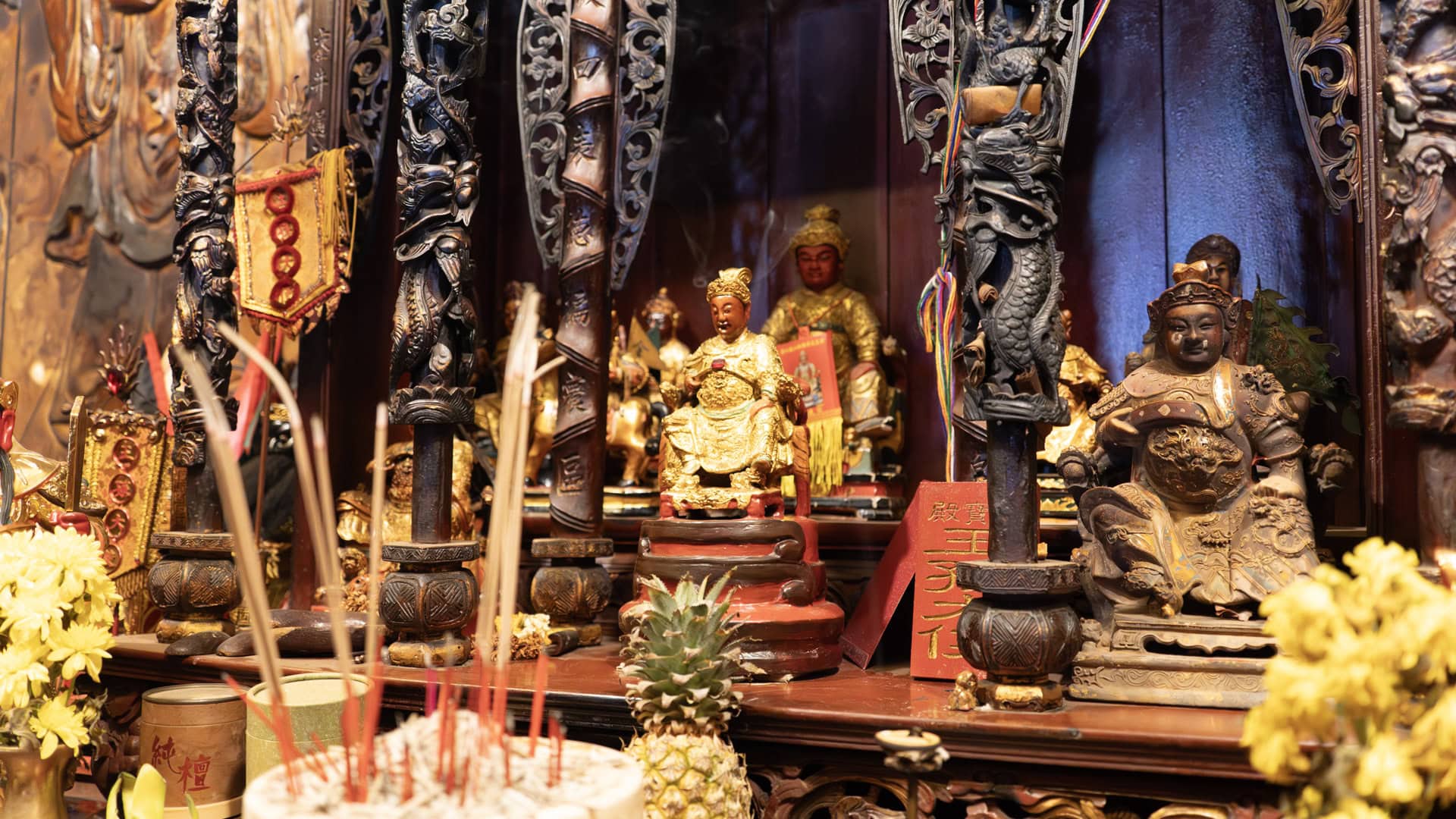 Small deity statues sit inside a carved wooden altar at San Chao Saengtham, with incense sticks, yellow flowers, and a pineapple in front.