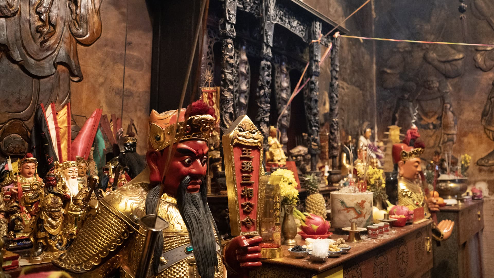 A red-faced guardian statue stands beside an altar inside San Chao Saengtham, with fruit offerings, incense, and large wall relief figures behind it.