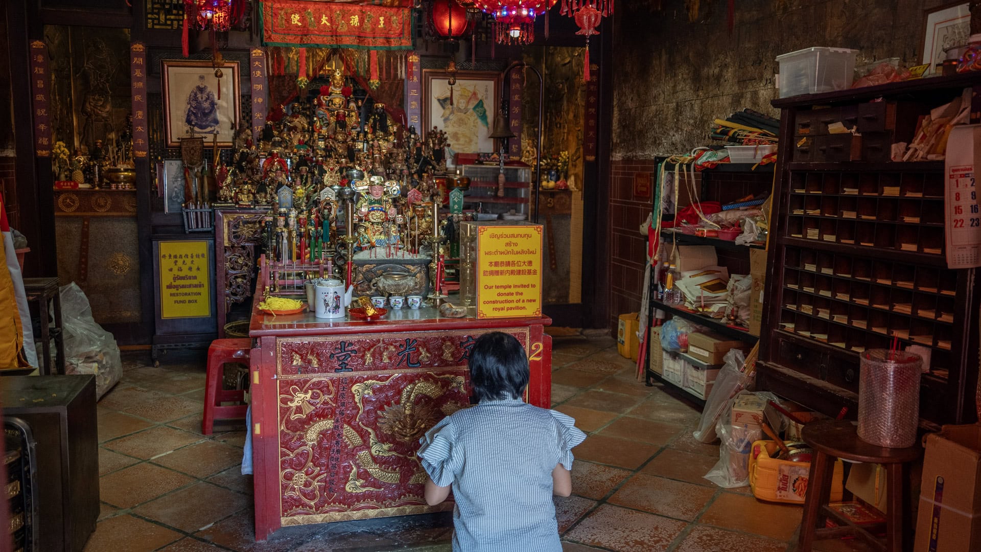 A person kneels before the main altar inside San Chao Saengtham, with many small deity figures, red lanterns, donation signs, and wooden storage shelves around the shrine table.