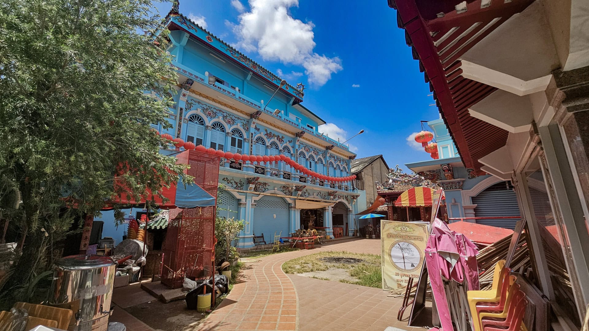 A curved brick path runs through the courtyard at San Chao Saengtham, with a blue building, red lanterns, and stacked chairs under a bright sky.