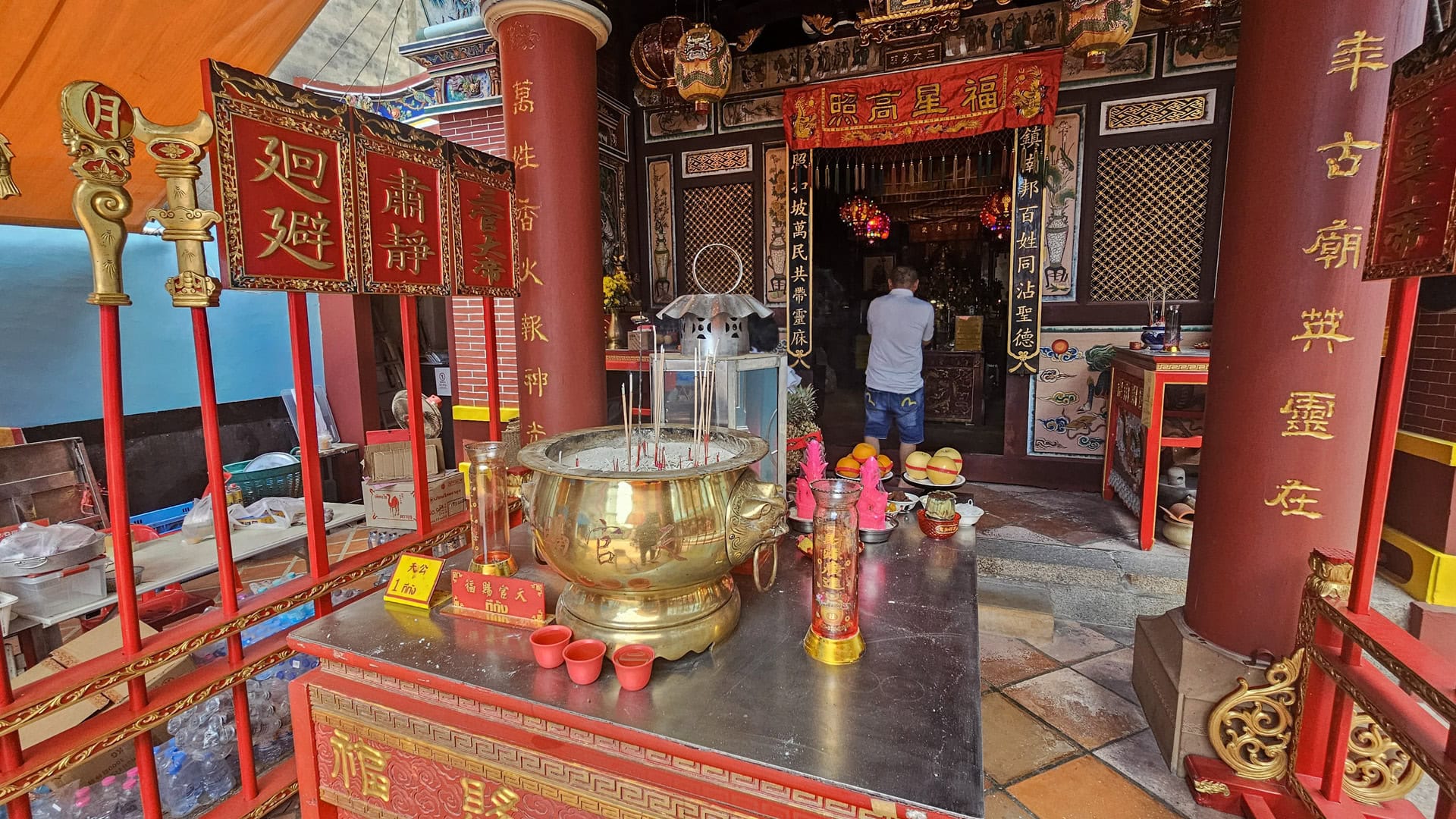 A brass incense burner sits on a metal-topped table between red pillars at San Chao Saengtham, with offerings and a person standing inside the hall beyond.