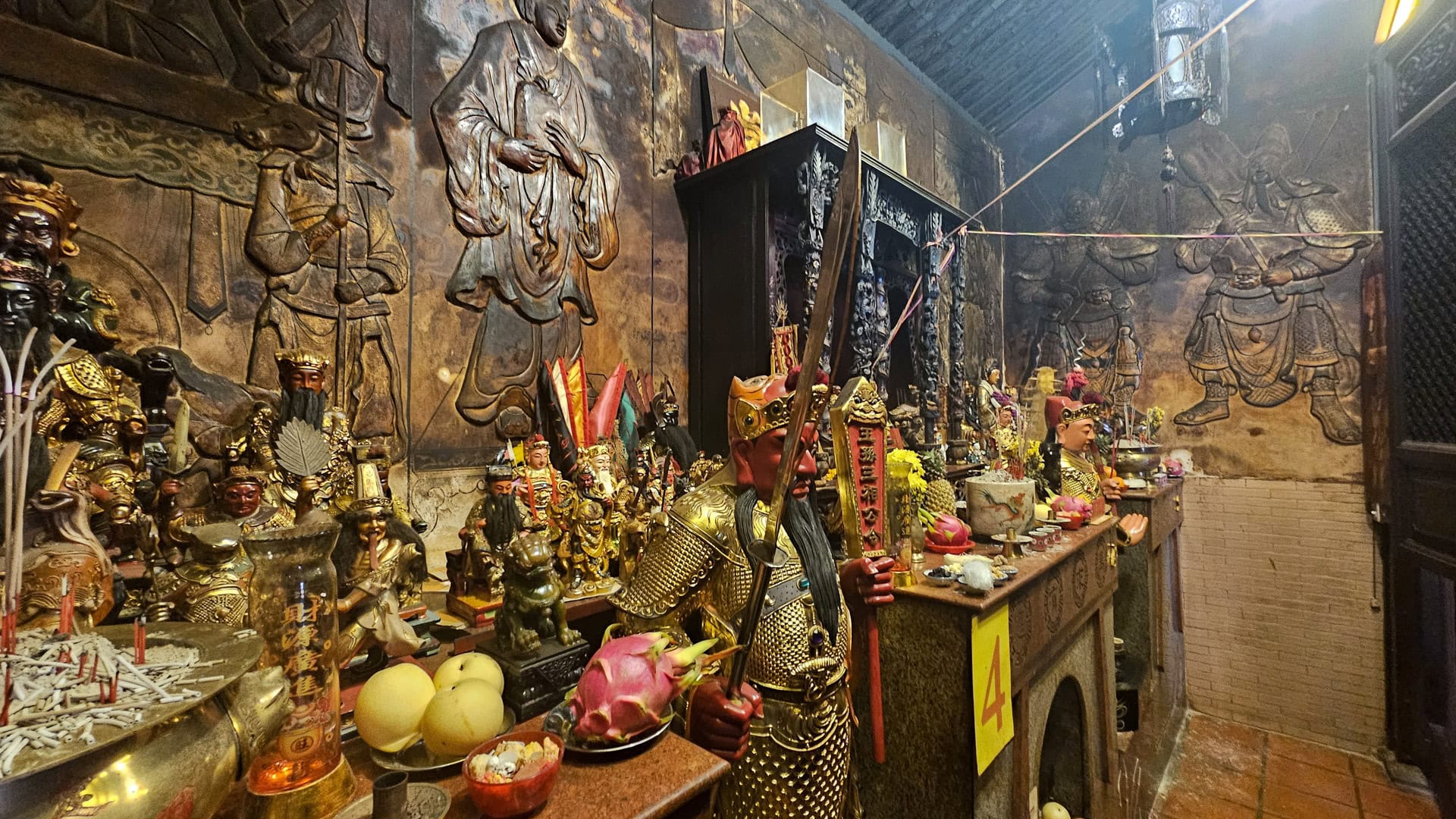 A red-faced guardian statue stands beside an altar inside San Chao Saengtham, with fruit offerings, incense, and large wall relief figures behind it.